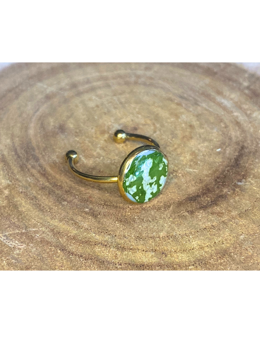 A gold-toned ring with a round bezel setting containing a green and white leaf, displayed on a rustic wood background.