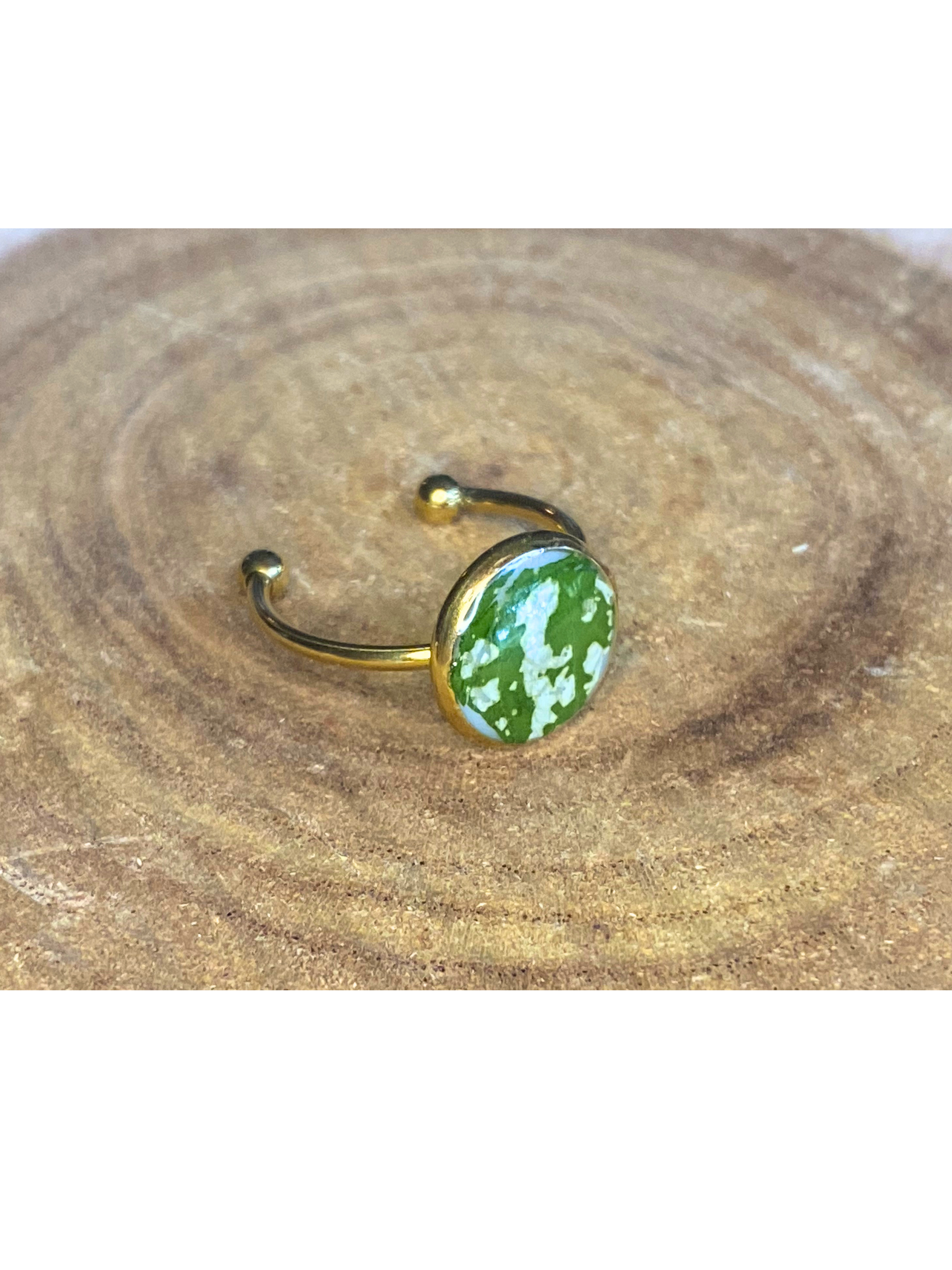 A gold-toned ring with a round bezel setting containing a green and white leaf, displayed on a rustic wood background.
