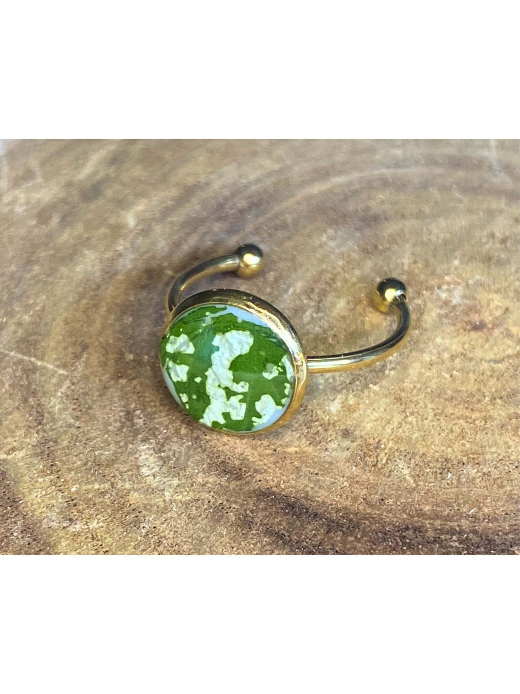 A gold-toned ring with a round bezel setting containing a green and white leaf, displayed on a rustic wood background.
