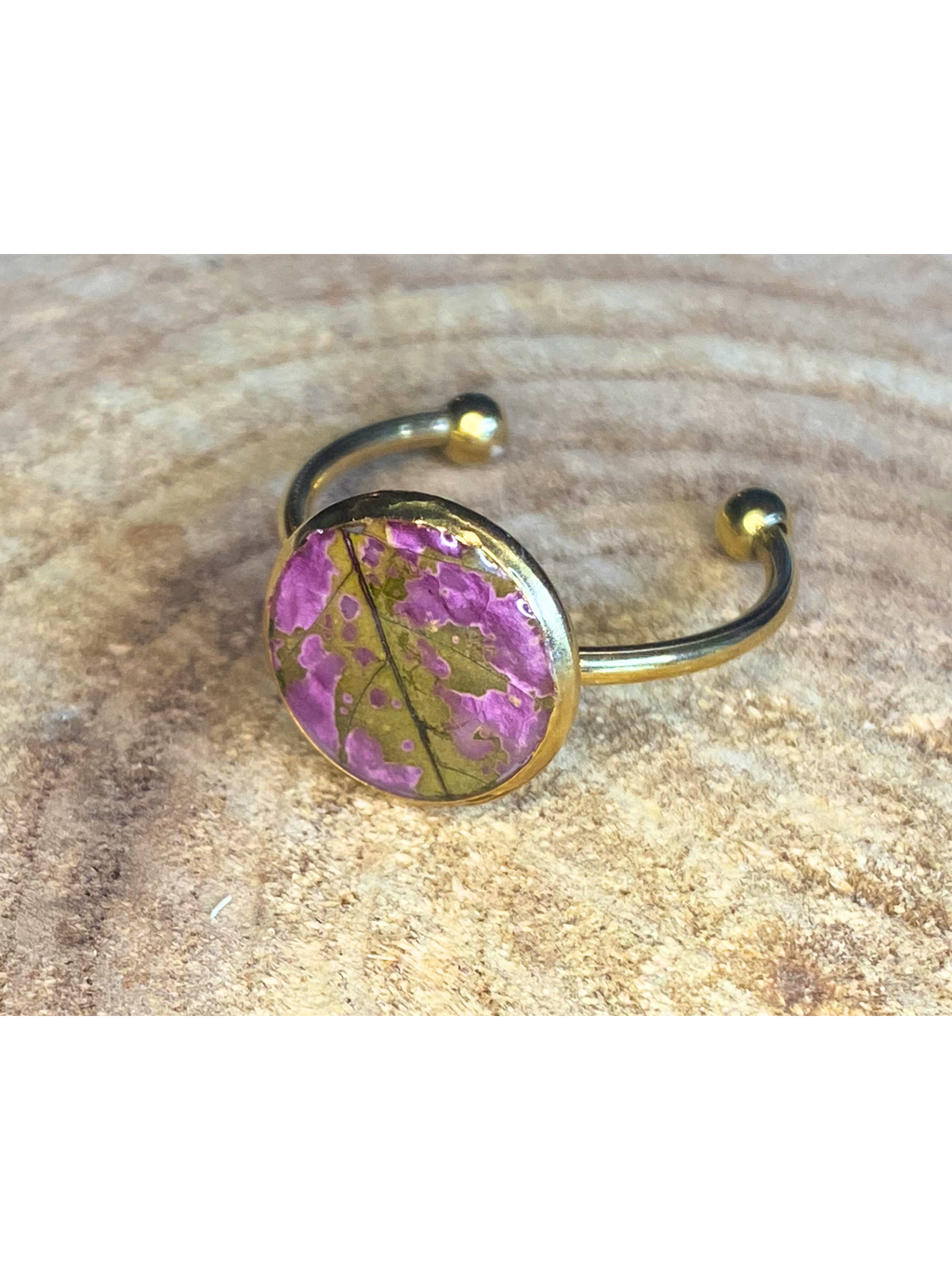 A silver-toned ring with a round bezel setting containing a pink and green leaf, displayed on a rustic wood background.