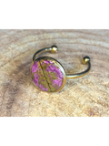A silver-toned ring with a round bezel setting containing a pink and green leaf, displayed on a rustic wood background.