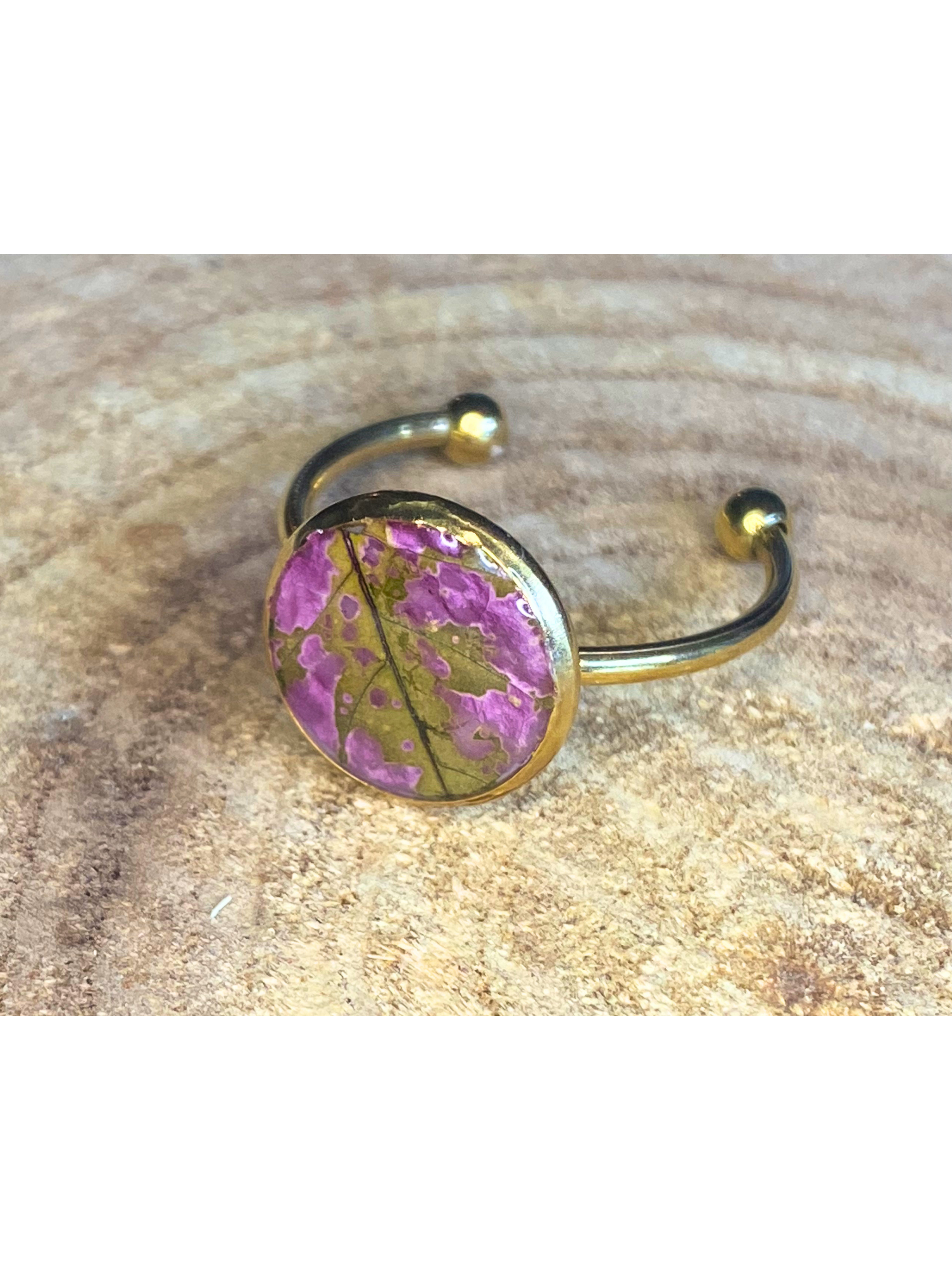 A silver-toned ring with a round bezel setting containing a pink and green leaf, displayed on a rustic wood background.