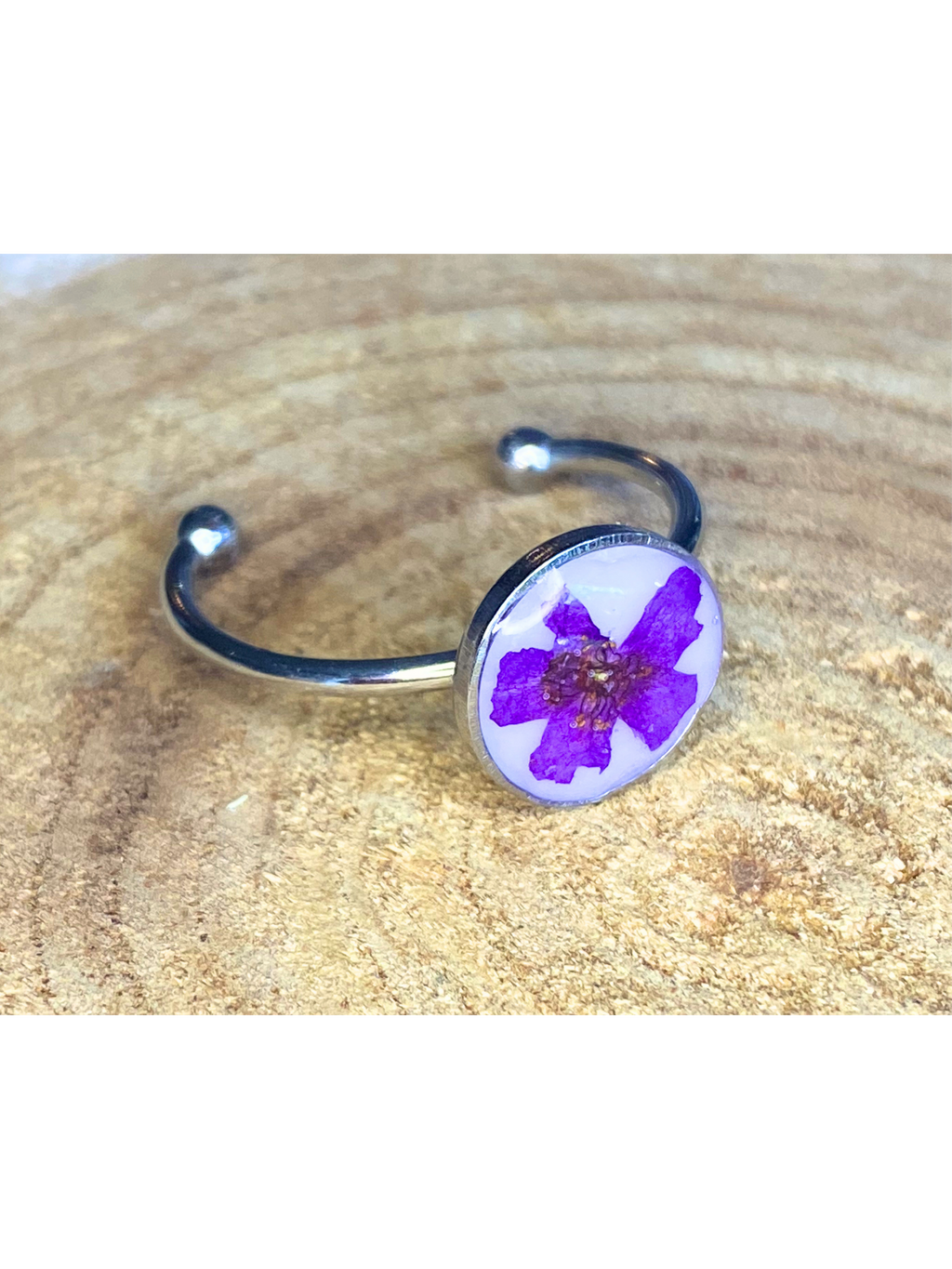 A silver-toned ring featuring a circular bezel with a pressed purple flower on a white background, displayed on a textured wood surface.