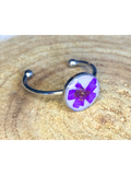 A silver-toned ring featuring a circular bezel with a pressed purple flower on a white background, displayed on a textured wood surface.