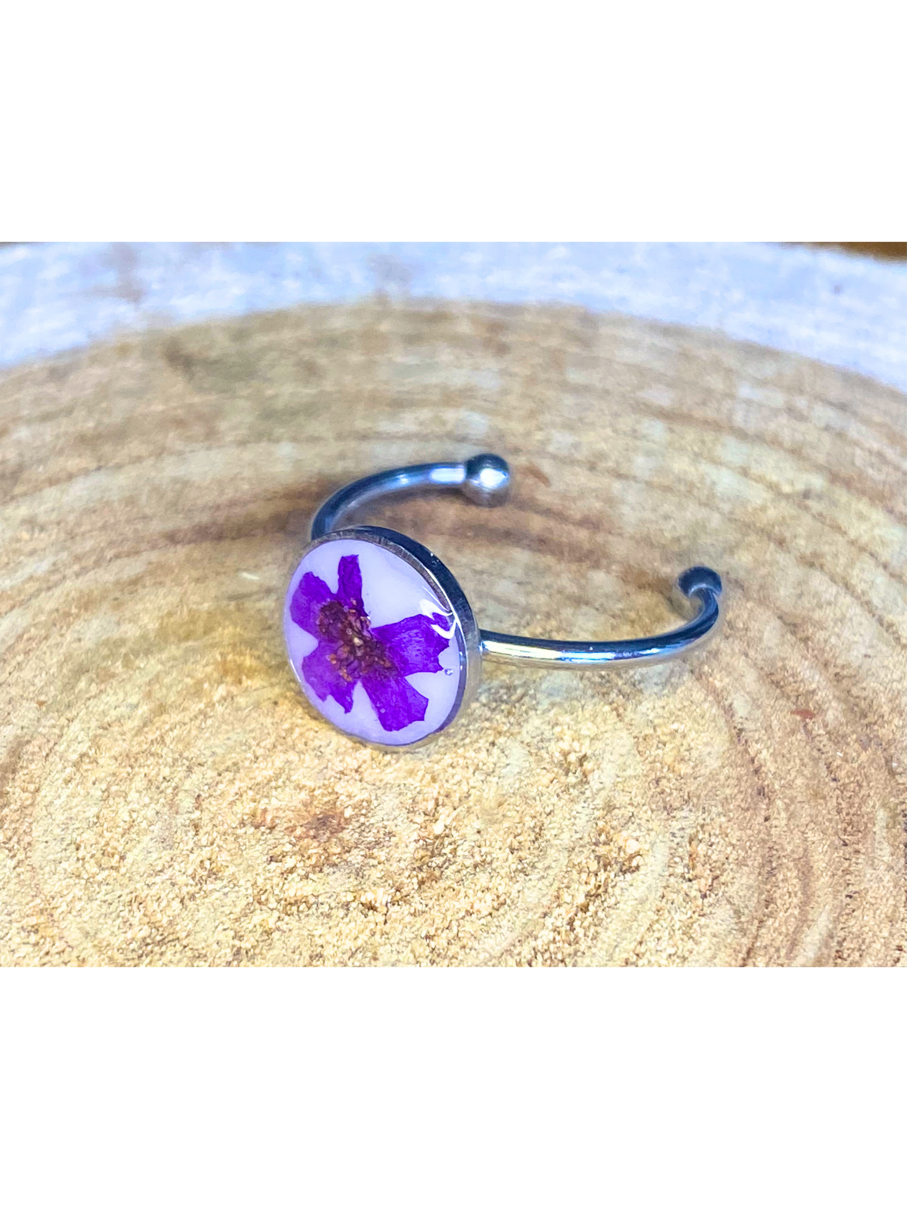 A silver-toned ring featuring a circular bezel with a pressed purple flower on a white background, displayed on a textured wood surface.