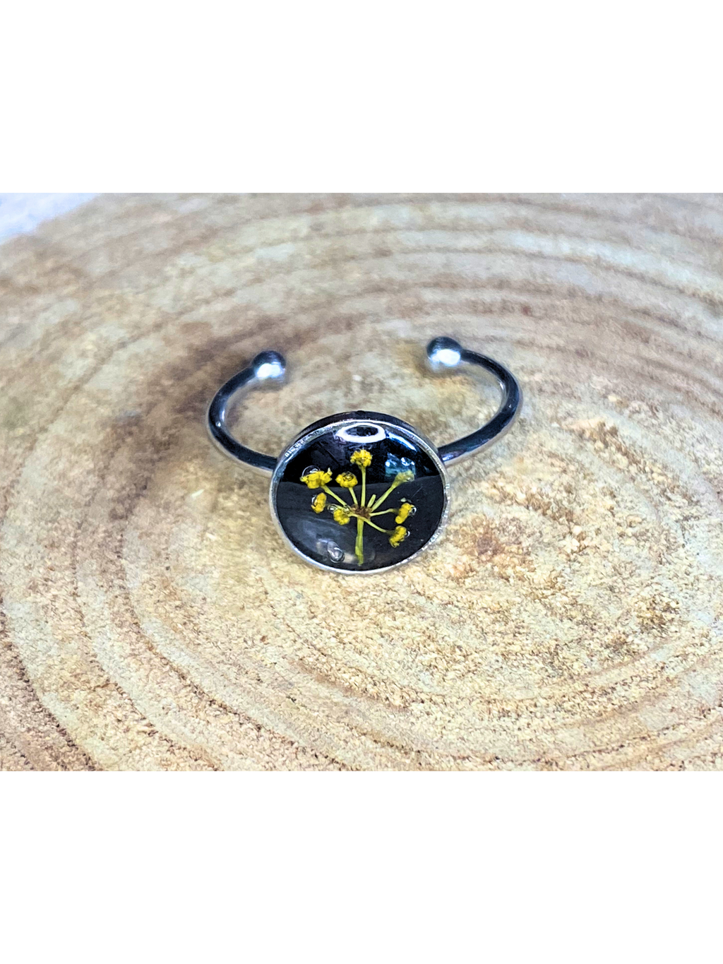 A silver-toned ring with a round bezel setting containing a yellow flower with a black background, displayed on a rustic wood background.