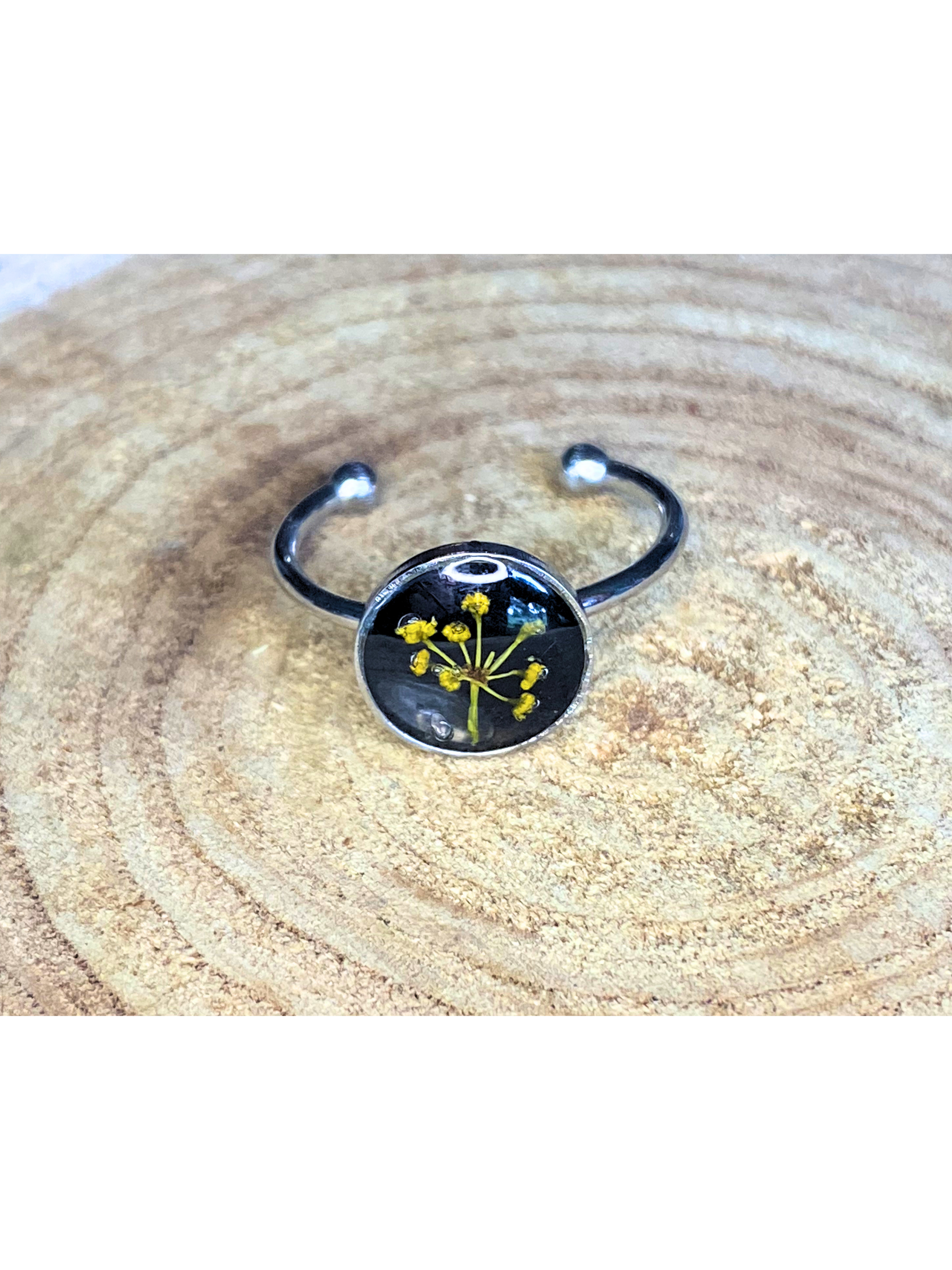 A silver-toned ring with a round bezel setting containing a yellow flower with a black background, displayed on a rustic wood background.
