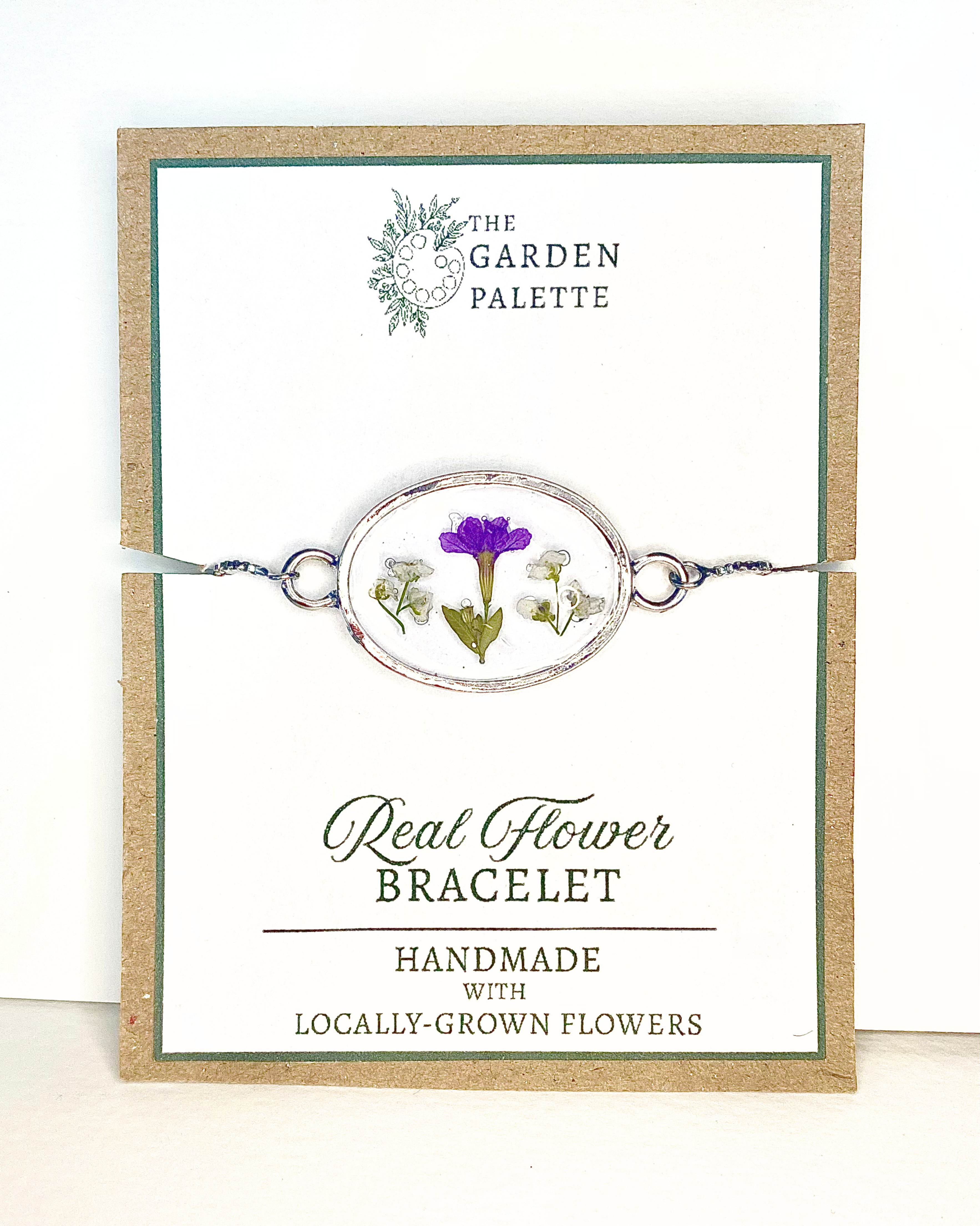 A silver bracelet with pressed purple and white flowers, displayed on a branded card that reads 'The Garden Palette' and 'Real Flower Bracelet'.