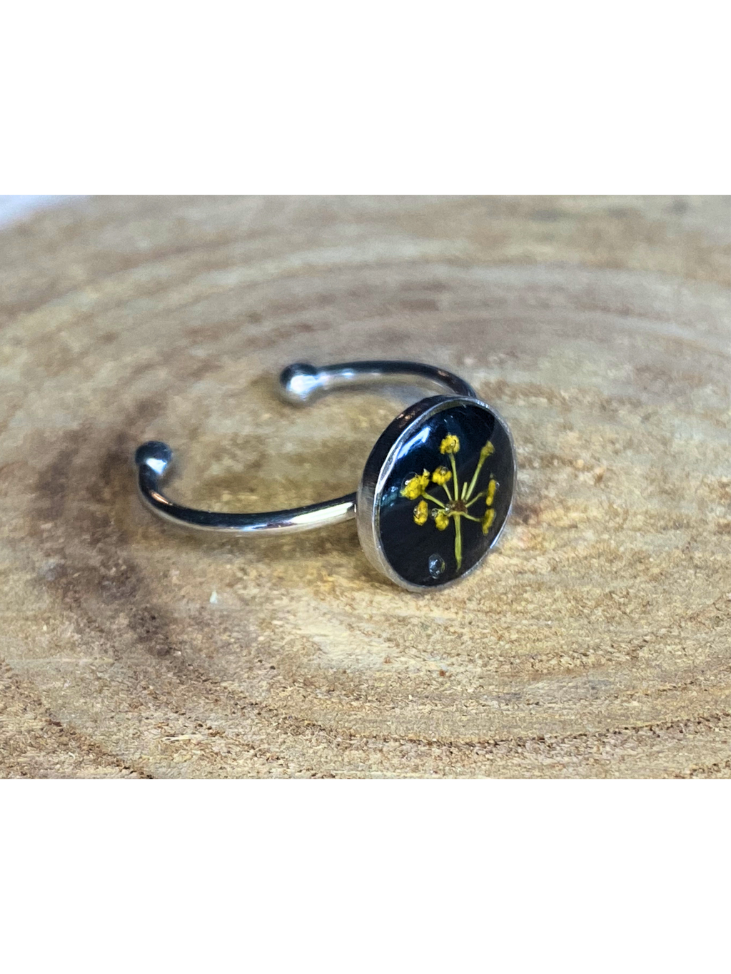 A silver-toned ring with a round bezel setting containing a yellow flower with a black background, displayed on a rustic wood background.