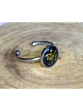 A silver-toned ring with a round bezel setting containing a yellow flower with a black background, displayed on a rustic wood background.