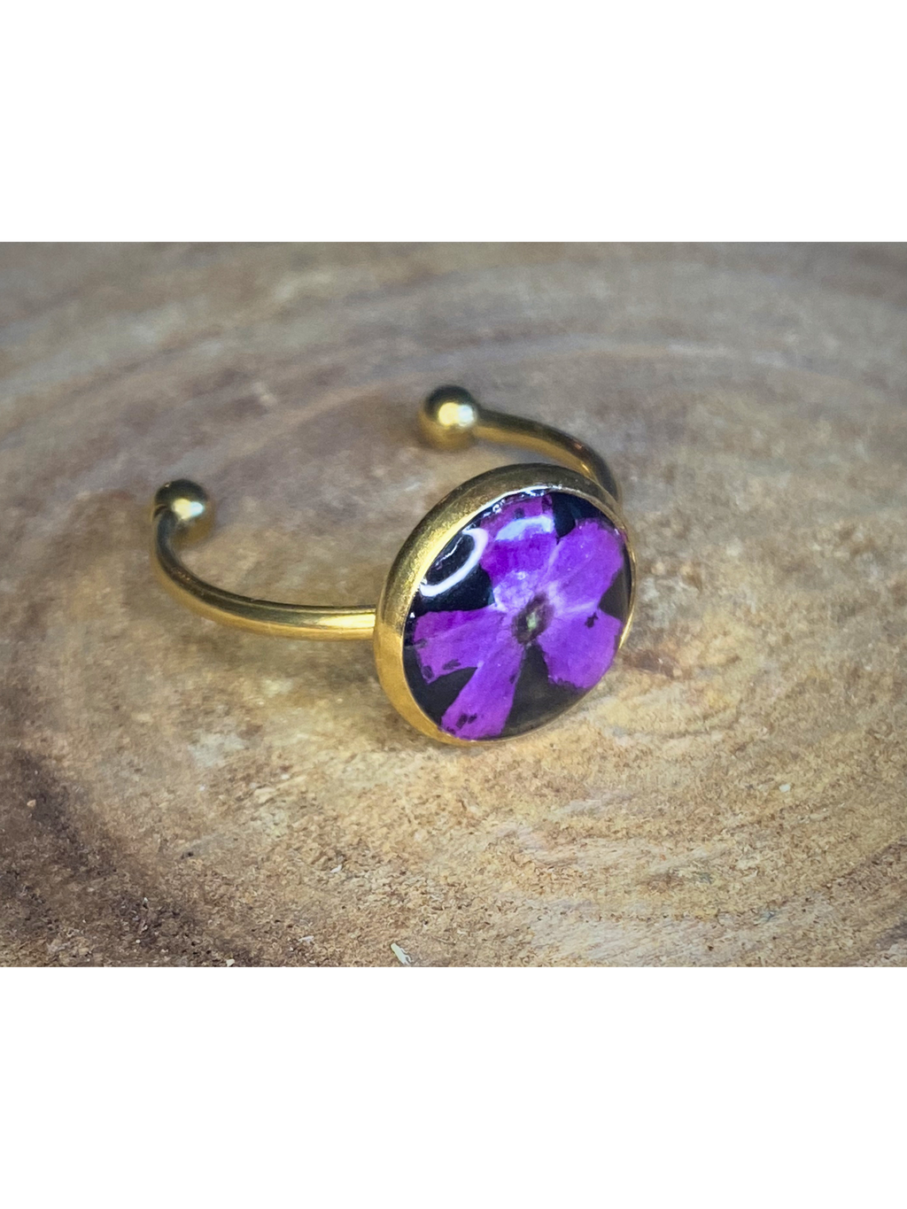 A gold-toned ring featuring a round bezel set with a purple flower on a black background, resting on a textured wood surface.