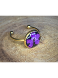 A gold-toned ring featuring a round bezel set with a purple flower on a black background, resting on a textured wood surface.