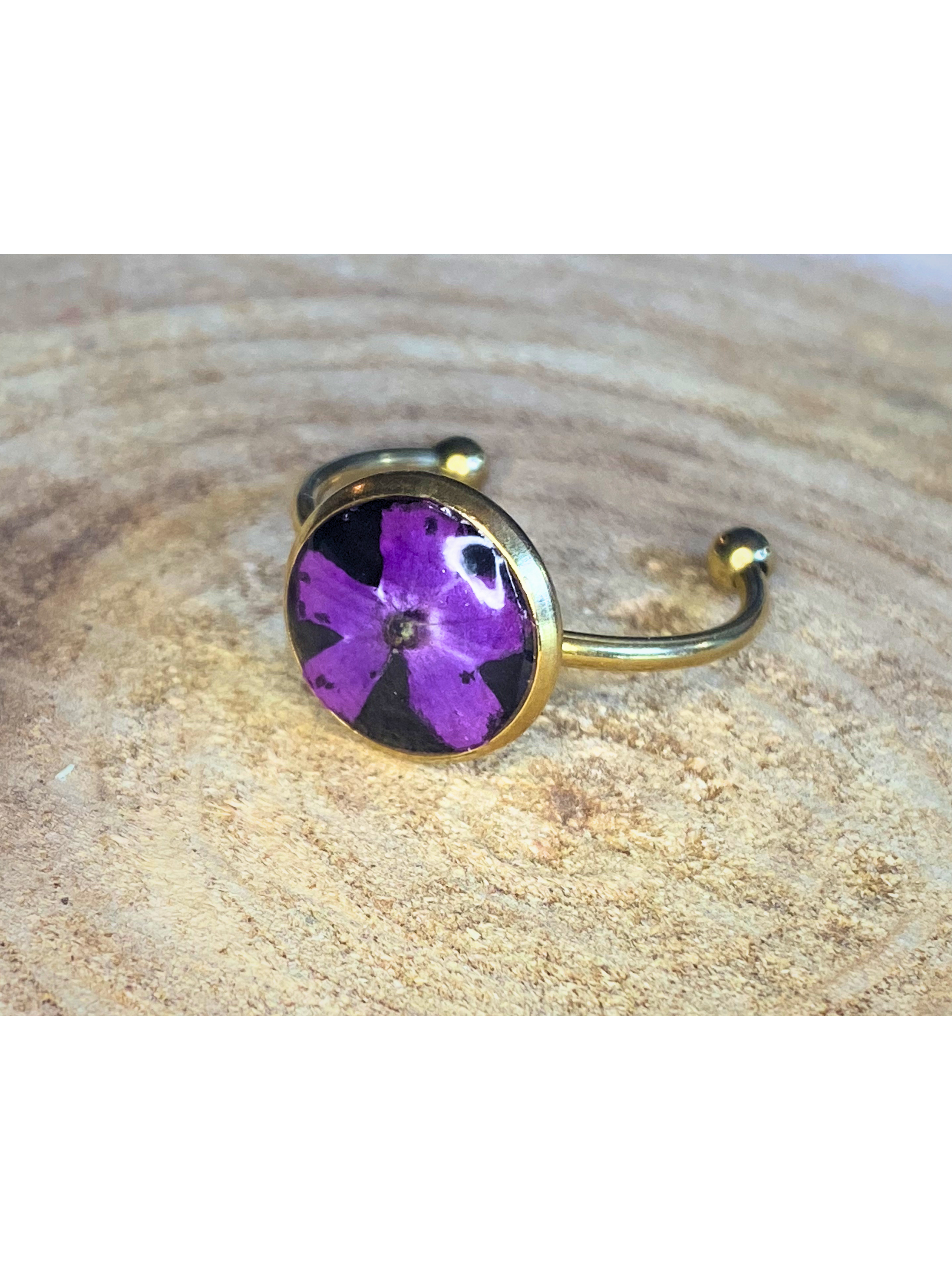 A gold-toned ring featuring a round bezel set with a purple flower on a black background, resting on a textured wood surface.