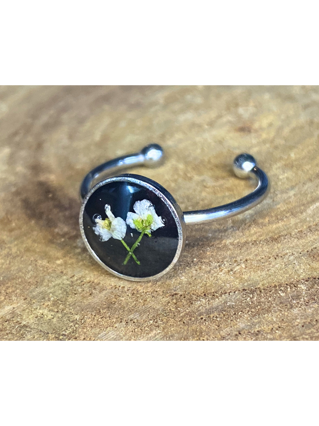 A silver-toned ring with a round bezel setting containing a white flower on a black background, displayed on a rustic wood surface.