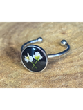A silver-toned ring with a round bezel setting containing a white flower on a black background, displayed on a rustic wood surface.