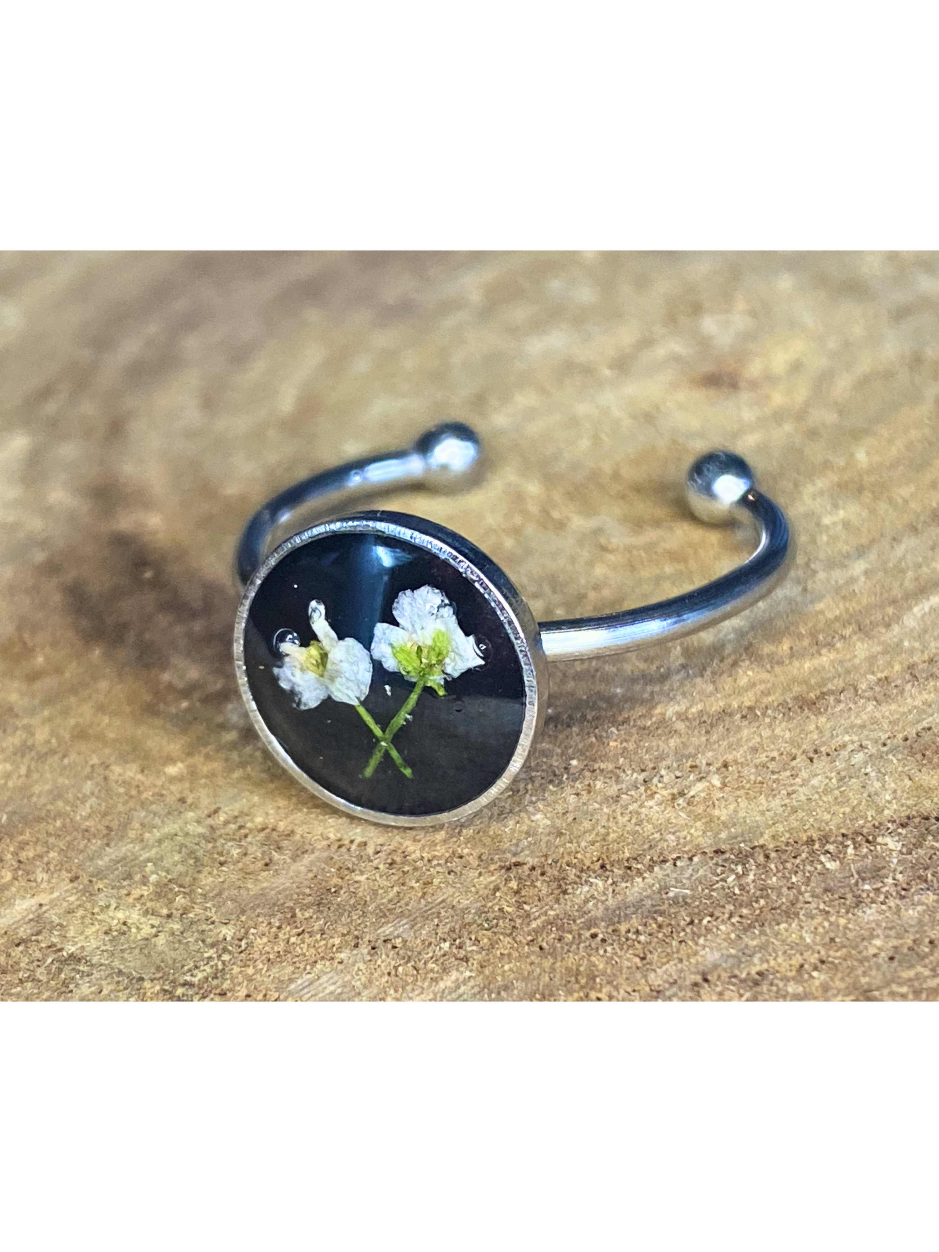 A silver-toned ring with a round bezel setting containing a white flower on a black background, displayed on a rustic wood surface.