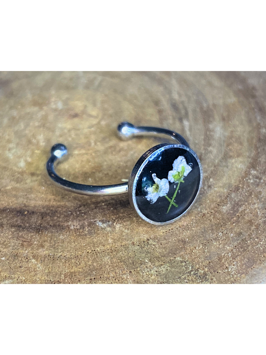 A silver-toned ring with a round bezel setting containing a white flower on a black background, displayed on a rustic wood surface.