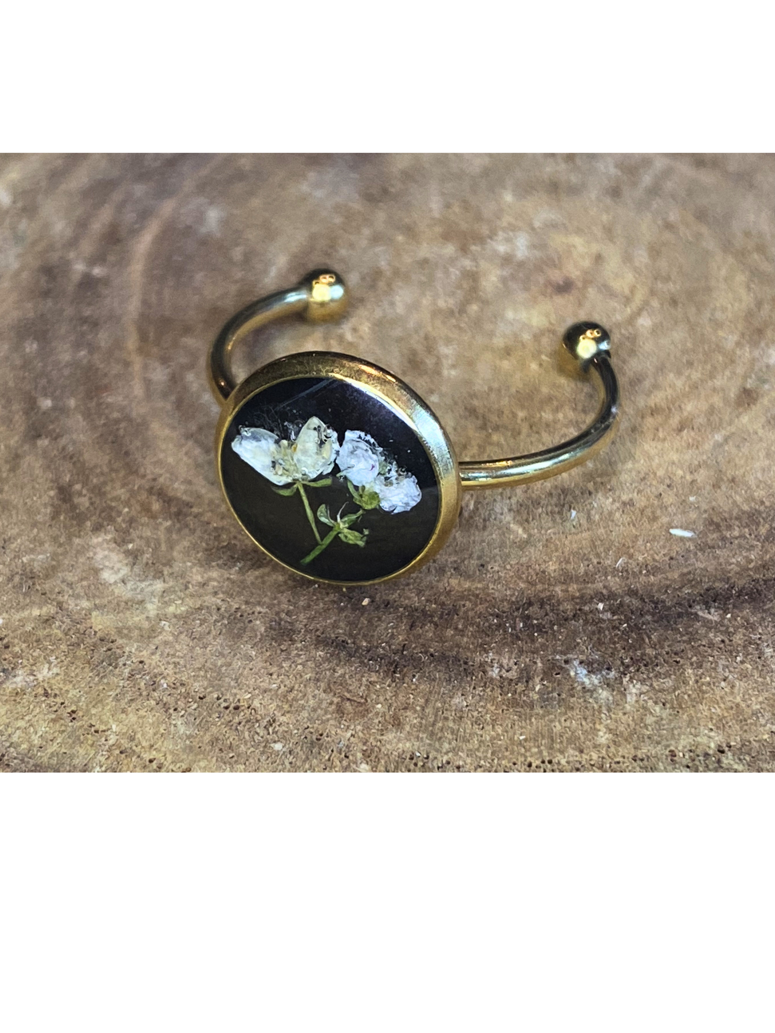 A gold-toned ring with a round bezel setting containing a white flower on a black background, displayed on a rustic wood surface.