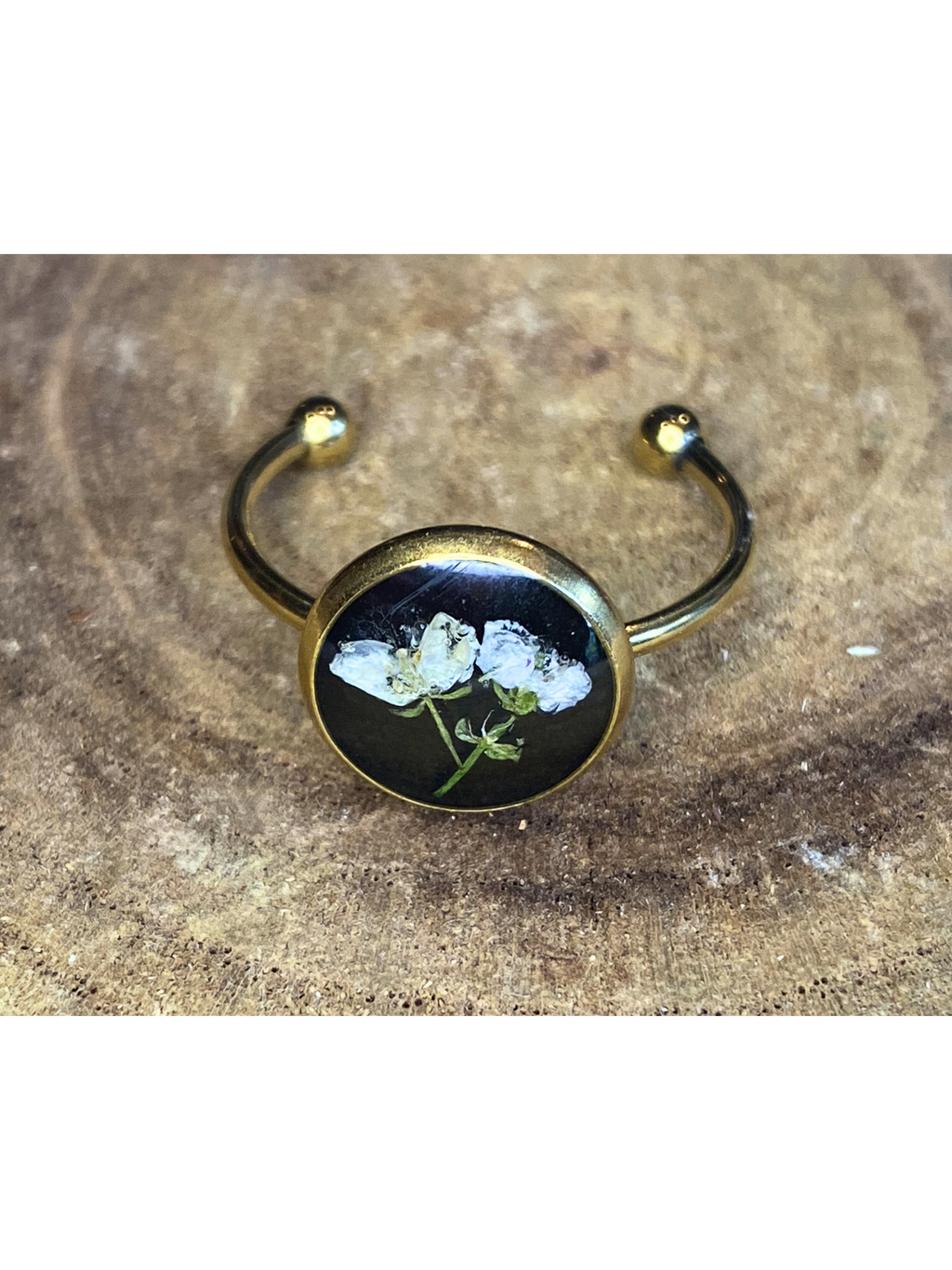 A gold-toned ring with a round bezel setting containing a white flower on a black background, displayed on a rustic wood surface.
