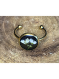A gold-toned ring with a round bezel setting containing a white flower on a black background, displayed on a rustic wood surface.