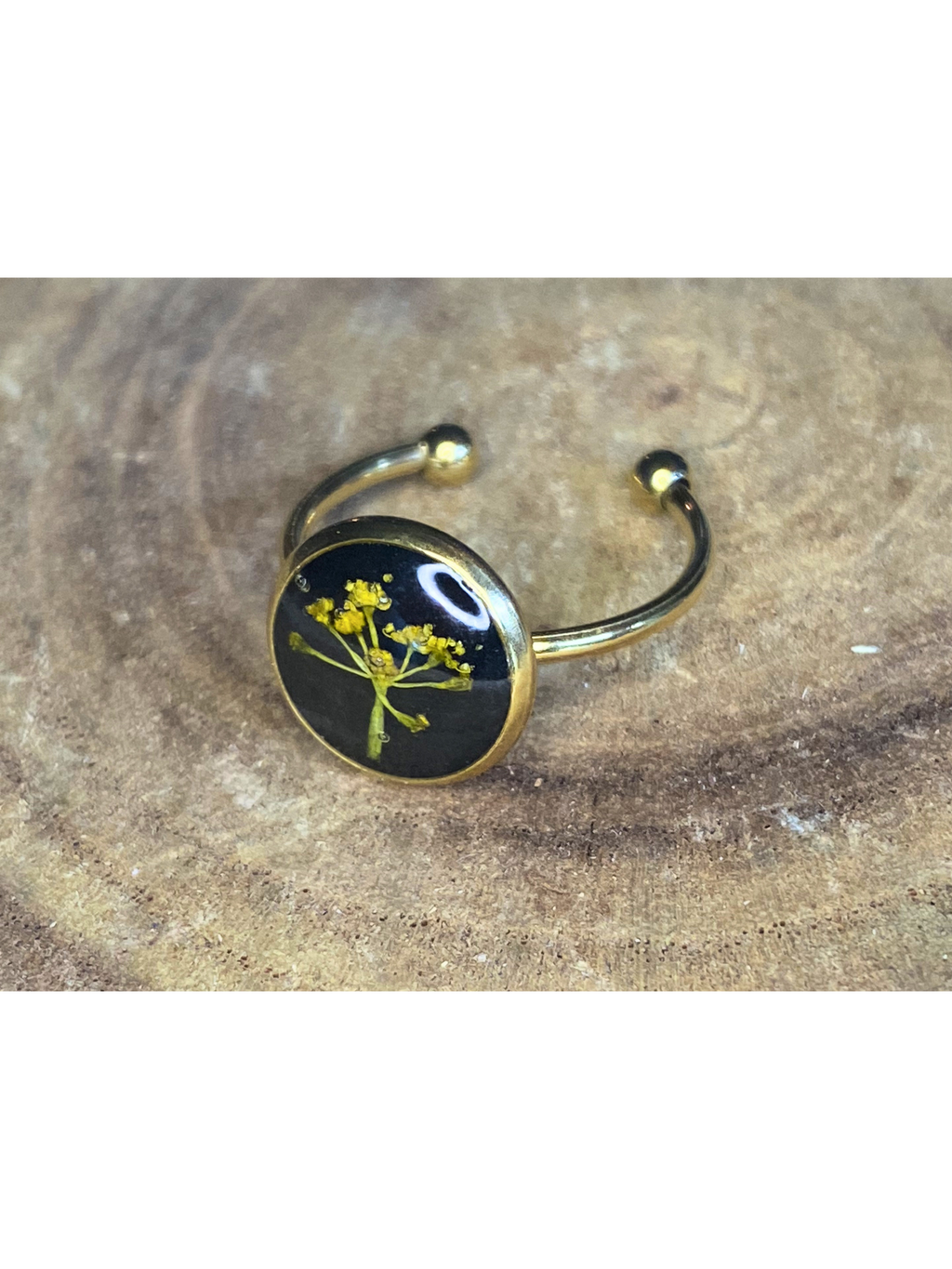A gold-toned ring with a round bezel setting containing a yellow flower on a black background, displayed on a rustic wood surface.