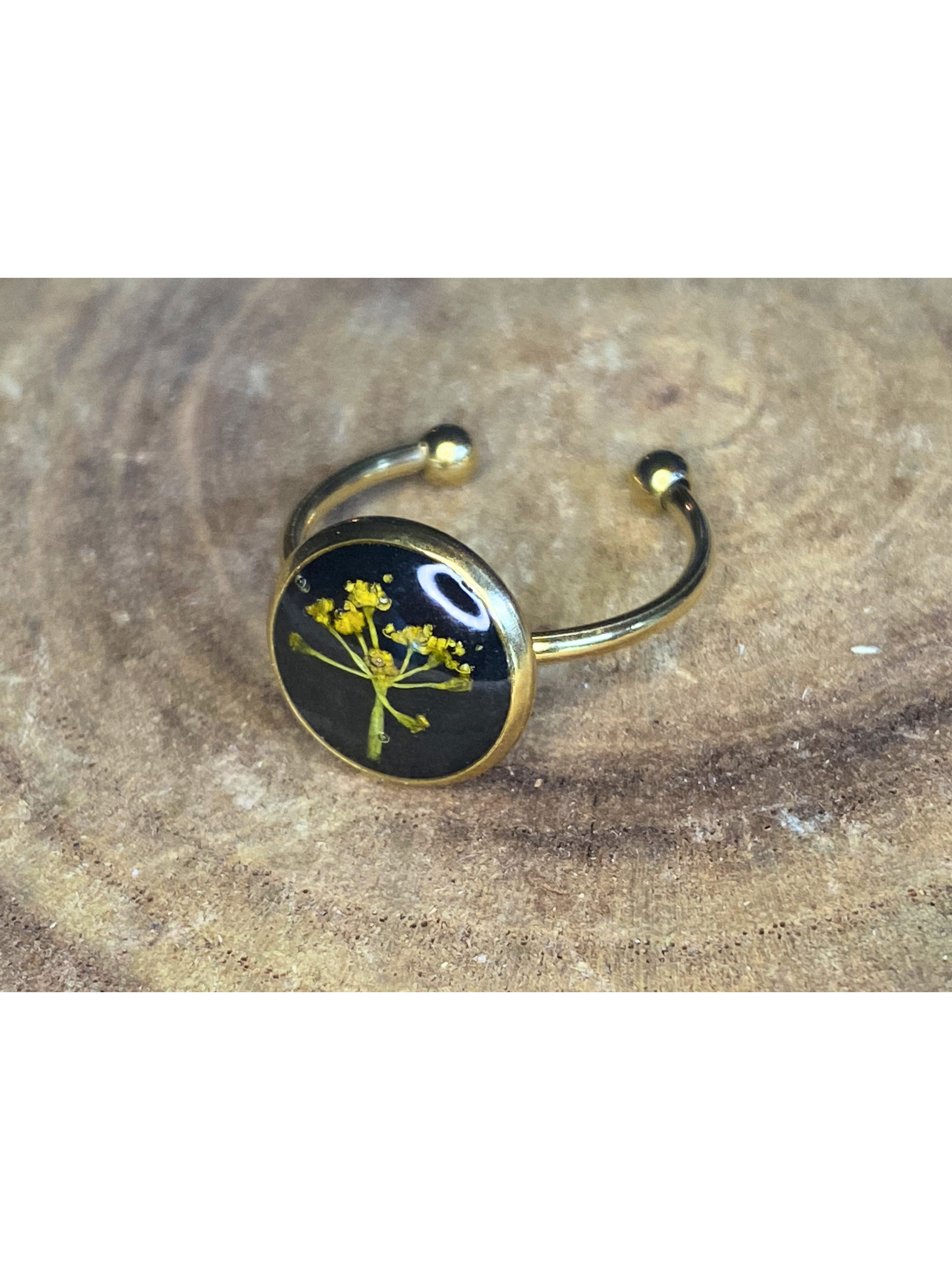 A gold-toned ring with a round bezel setting containing a yellow flower on a black background, displayed on a rustic wood surface.