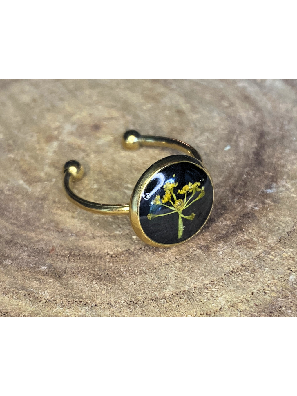 A gold-toned ring with a round bezel setting containing a yellow flower on a black background, displayed on a rustic wood surface.