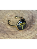 A gold-toned ring with a round bezel setting containing a yellow flower on a black background, displayed on a rustic wood surface.