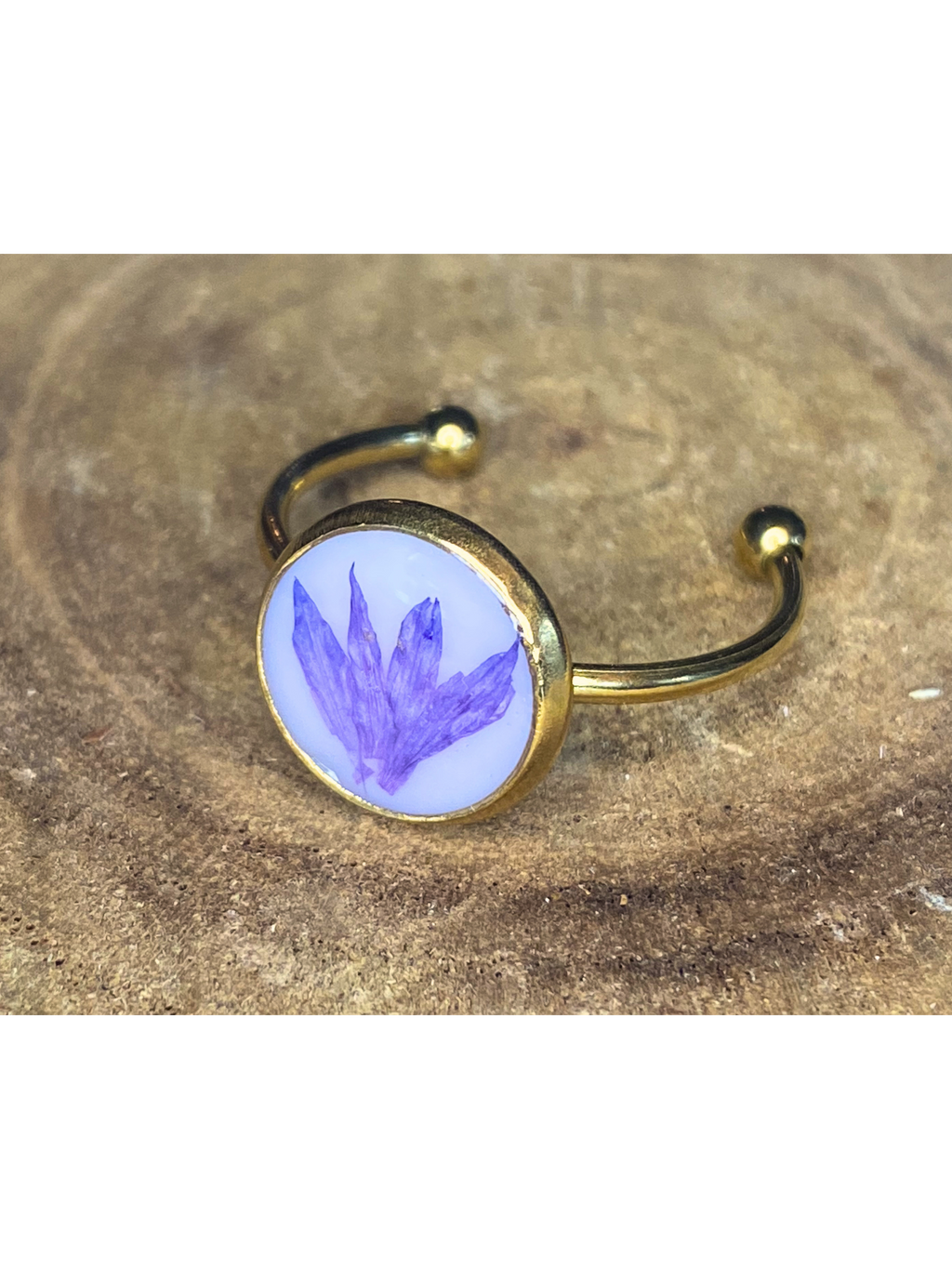 A gold-toned ring with a round bezel setting containing a blueish-purple flower petal on a white background, displayed on a rustic wood surface.