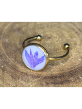 A gold-toned ring with a round bezel setting containing a blueish-purple flower petal on a white background, displayed on a rustic wood surface.
