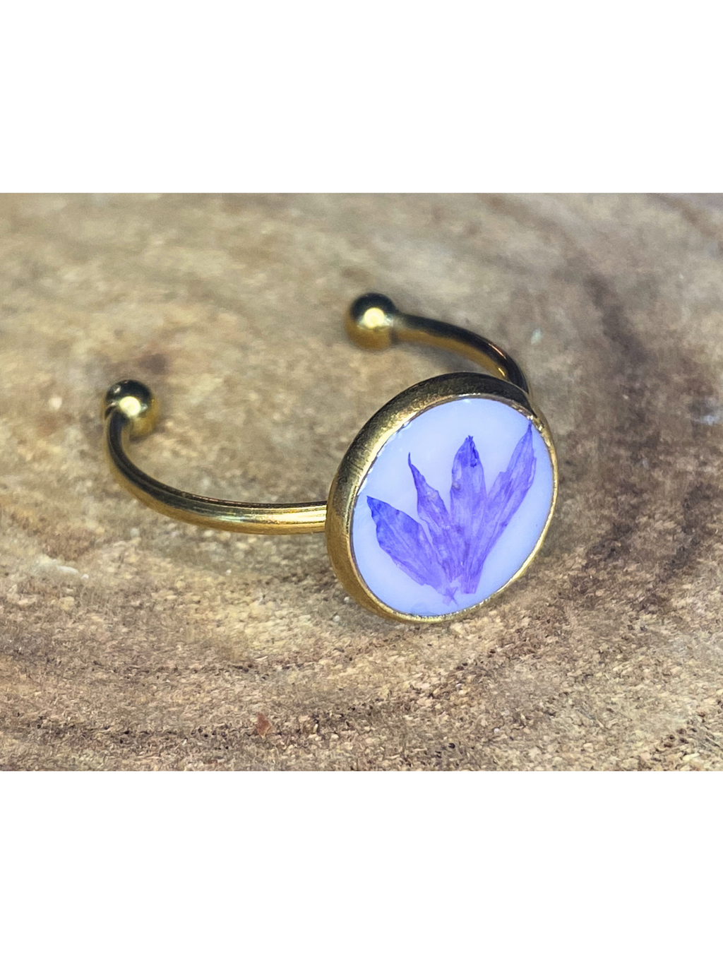 A gold-toned ring with a round bezel setting containing a blueish-purple flower petal on a white background, displayed on a rustic wood surface.