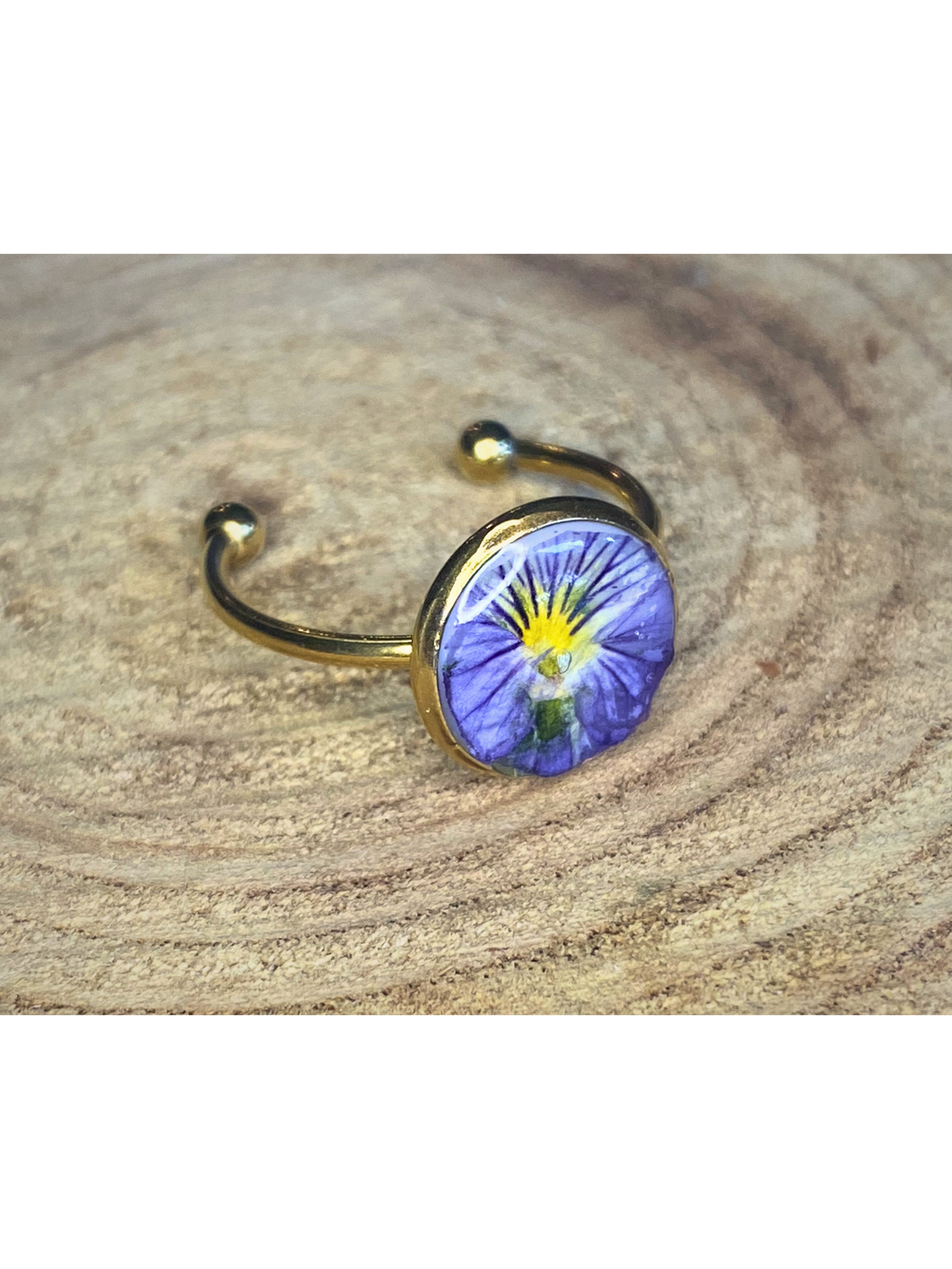 A gold-toned ring with a round bezel setting containing a blue and yellow flower, displayed on a rustic wood surface.