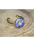 A gold-toned ring with a round bezel setting containing a blue and yellow flower, displayed on a rustic wood surface.