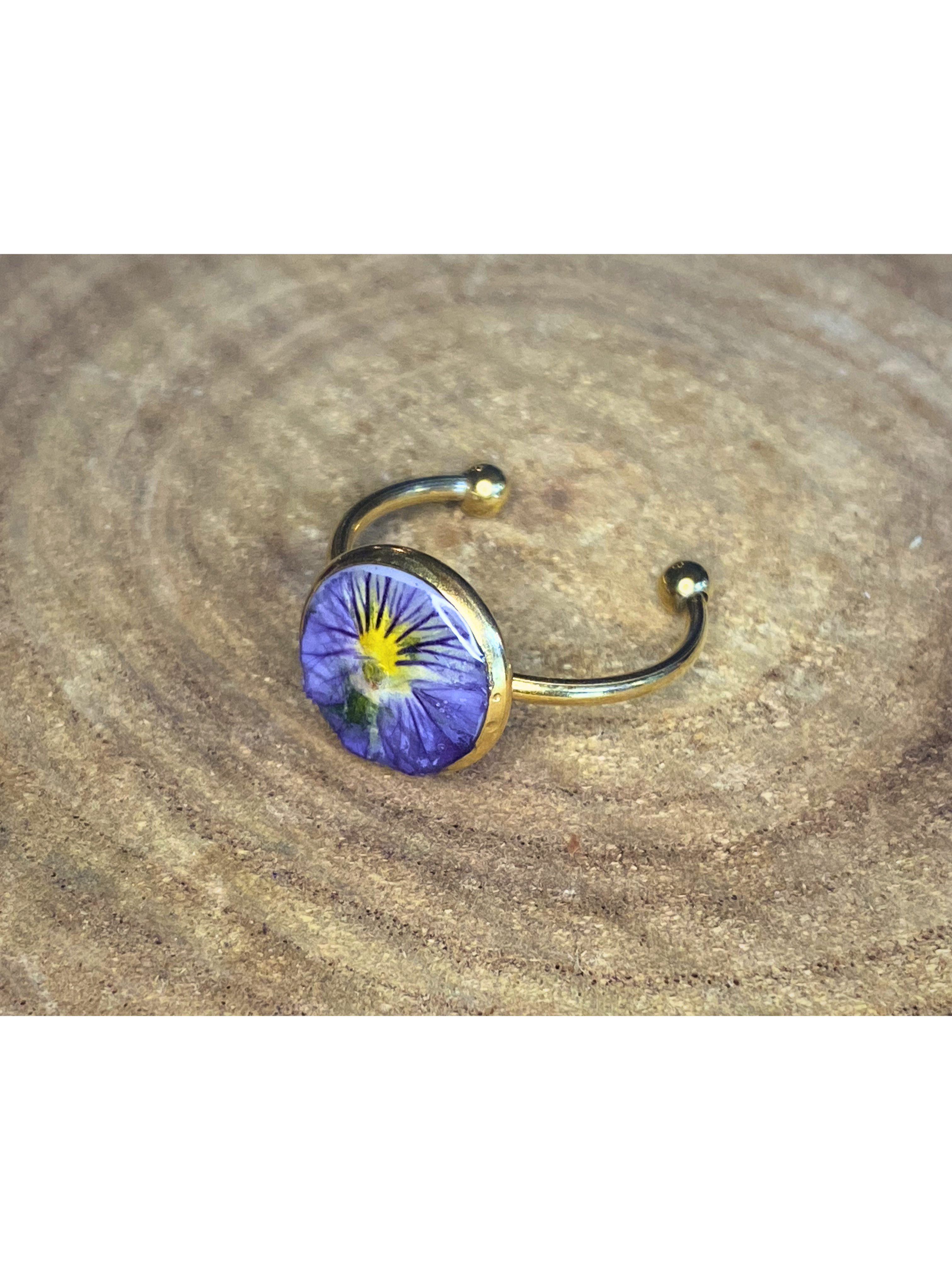 A gold-toned ring with a round bezel setting containing a blue and yellow flower, displayed on a rustic wood surface.
