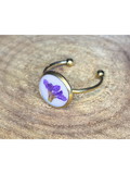 A gold-toned ring with a round bezel setting containing a purple flower on a white background, displayed on a rustic wood surface.