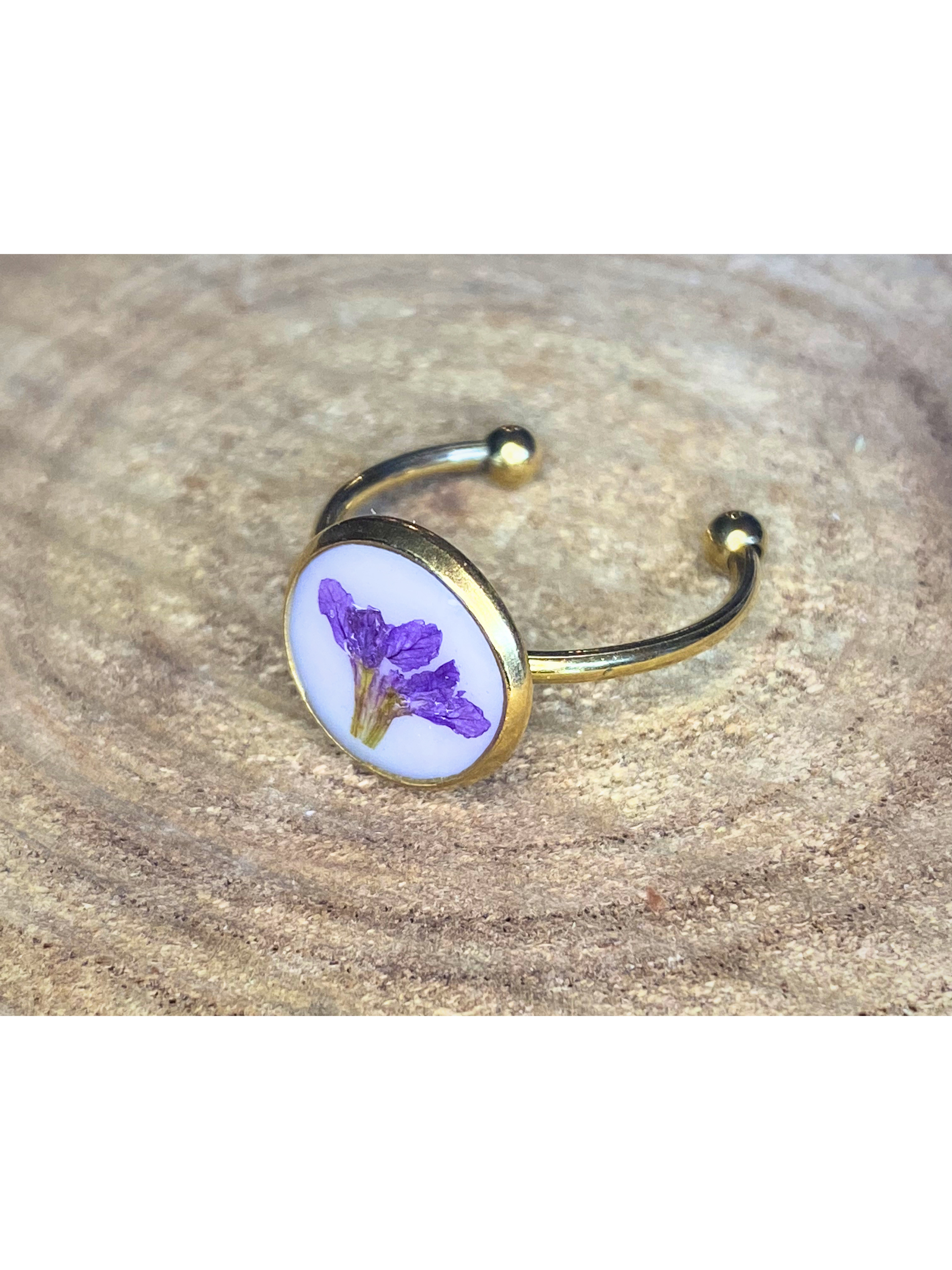 A gold-toned ring with a round bezel setting containing a purple flower on a white background, displayed on a rustic wood surface.