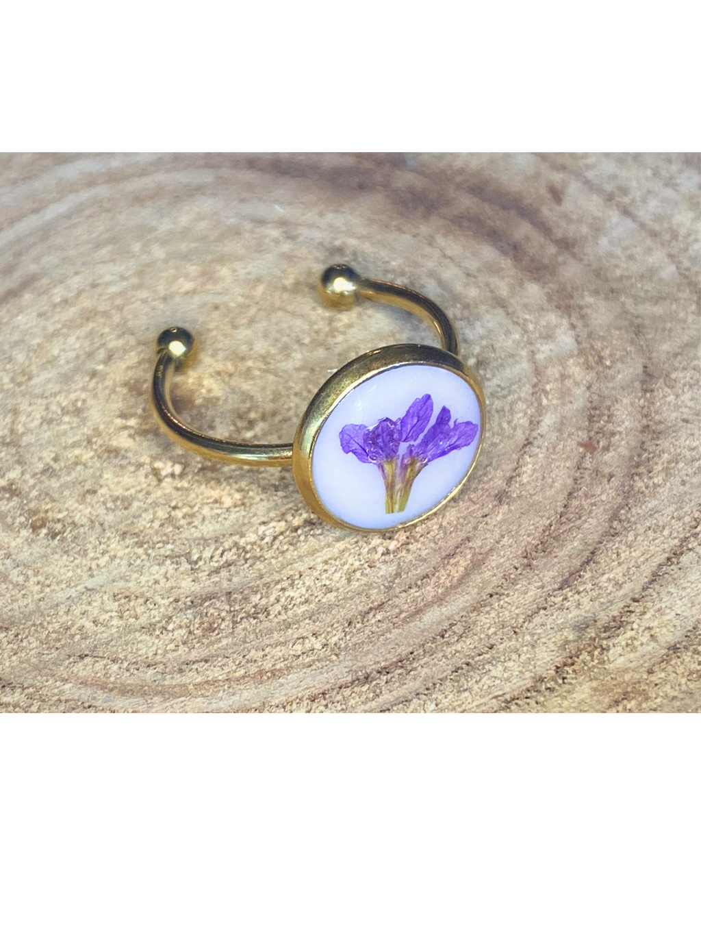 A gold-toned ring with a round bezel setting containing a purple flower on a white background, displayed on a rustic wood surface.