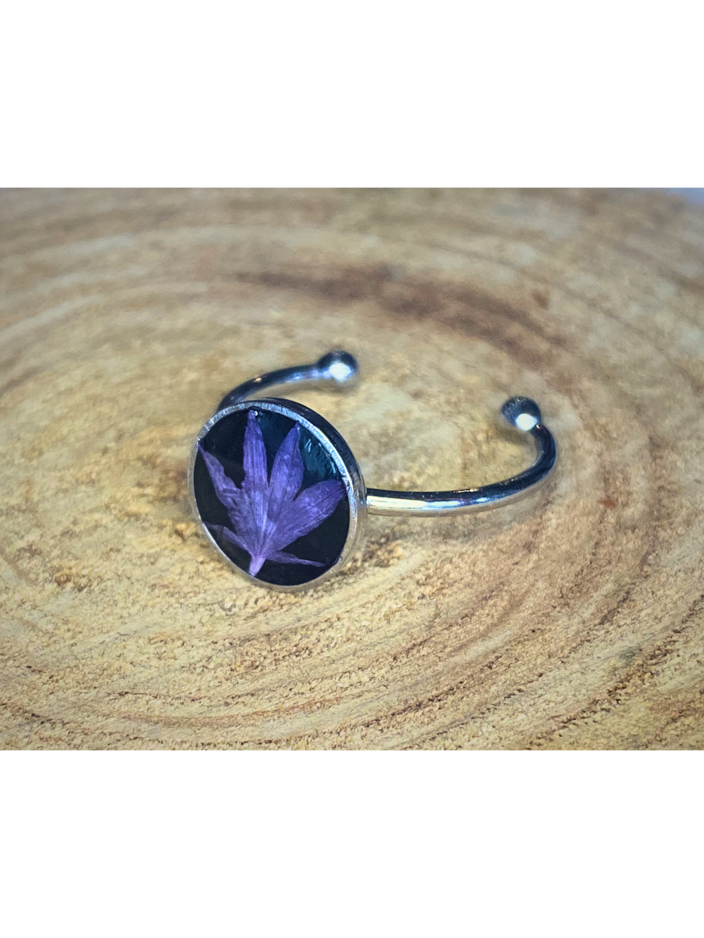 A silver-toned ring with a round bezel setting containing a blue flower petal on a black background, displayed on a rustic wood surface.