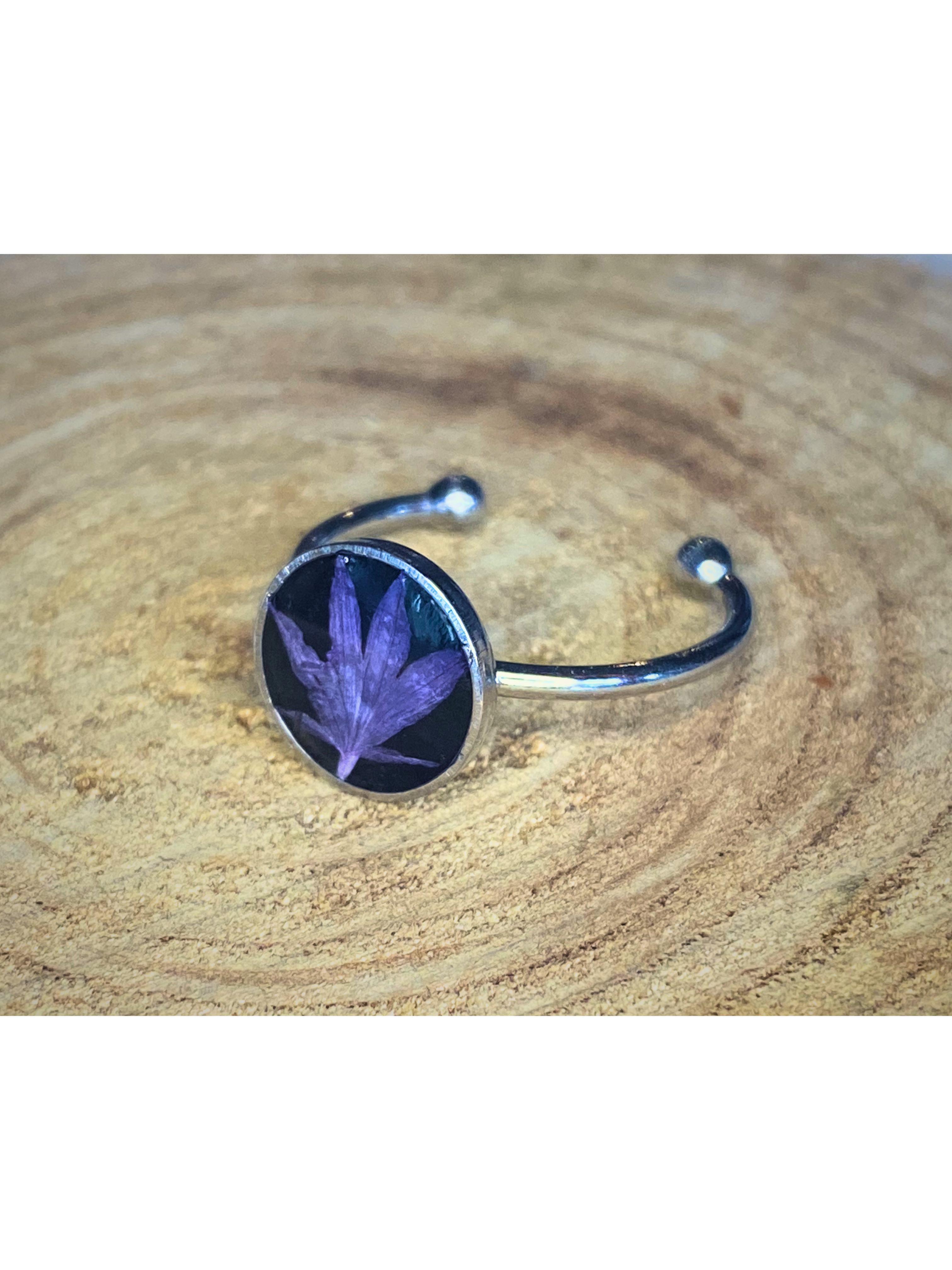 A silver-toned ring with a round bezel setting containing a blue flower petal on a black background, displayed on a rustic wood surface.