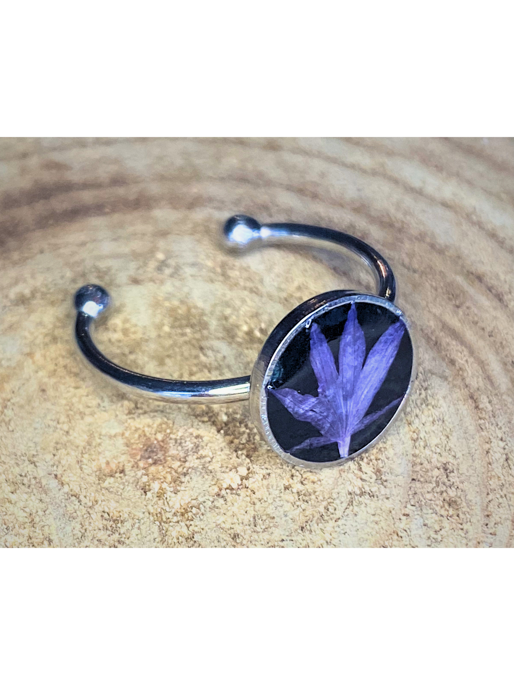 A silver-toned ring with a round bezel setting containing a blue flower petal on a black background, displayed on a rustic wood surface.
