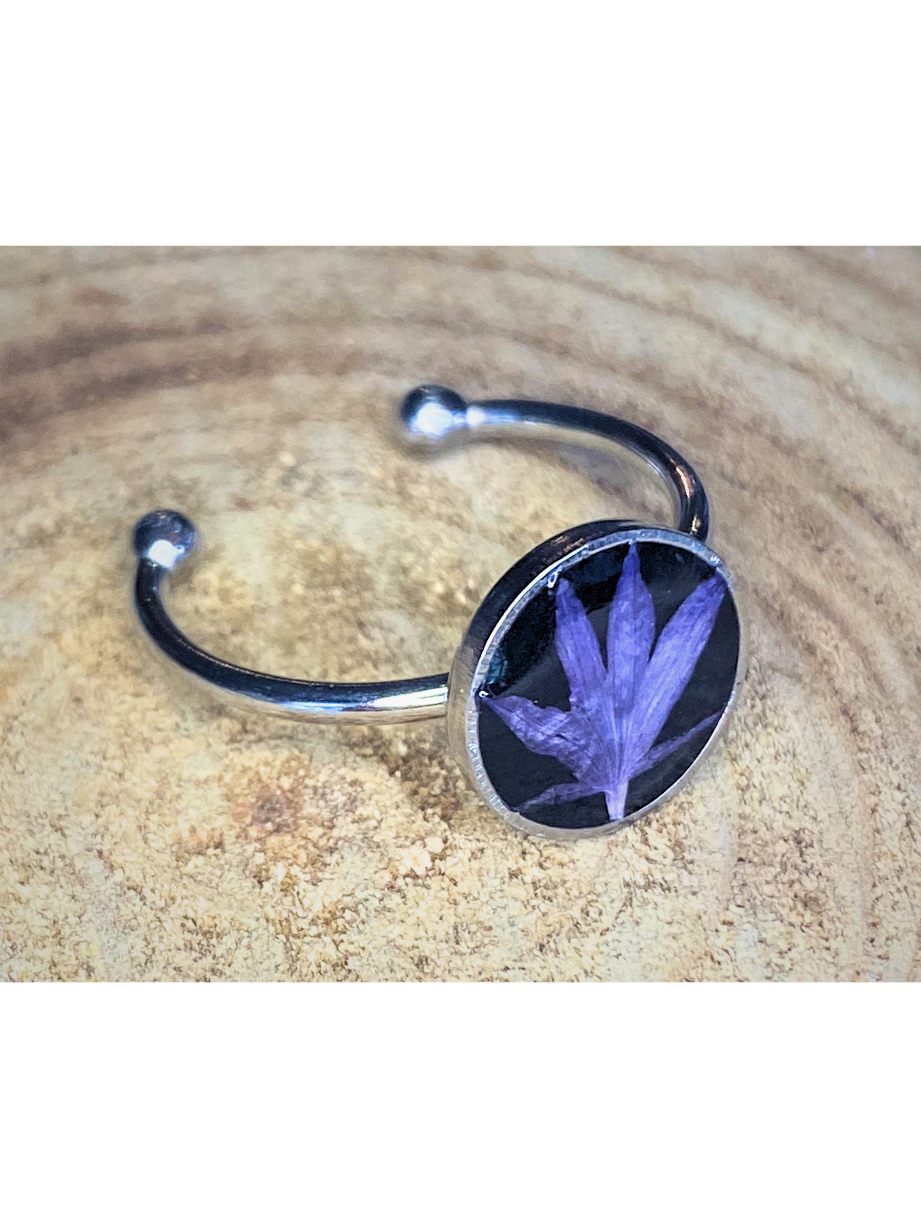 A silver-toned ring with a round bezel setting containing a blue flower petal on a black background, displayed on a rustic wood surface.