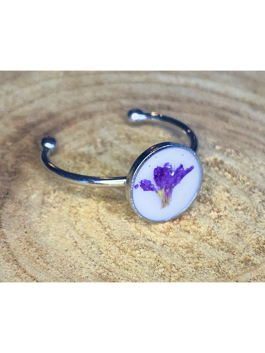 A silver-toned ring with a round bezel setting containing a purple flower on a white background, displayed on a rustic wood surface.