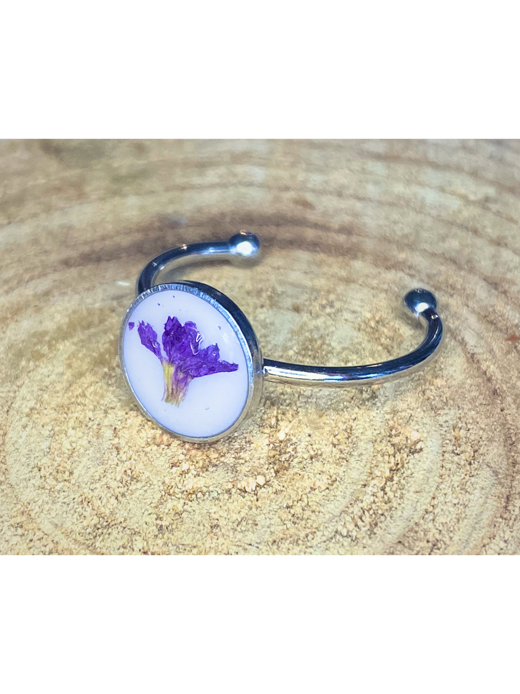 A silver-toned ring with a round bezel setting containing a purple flower on a white background, displayed on a rustic wood surface.