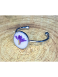 A silver-toned ring with a round bezel setting containing a purple flower on a white background, displayed on a rustic wood surface.