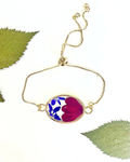 A delicate gold chain bracelet with an oval pendant containing pressed flower petals in blue, white, and red, displayed on a white background with decorative green leaves.