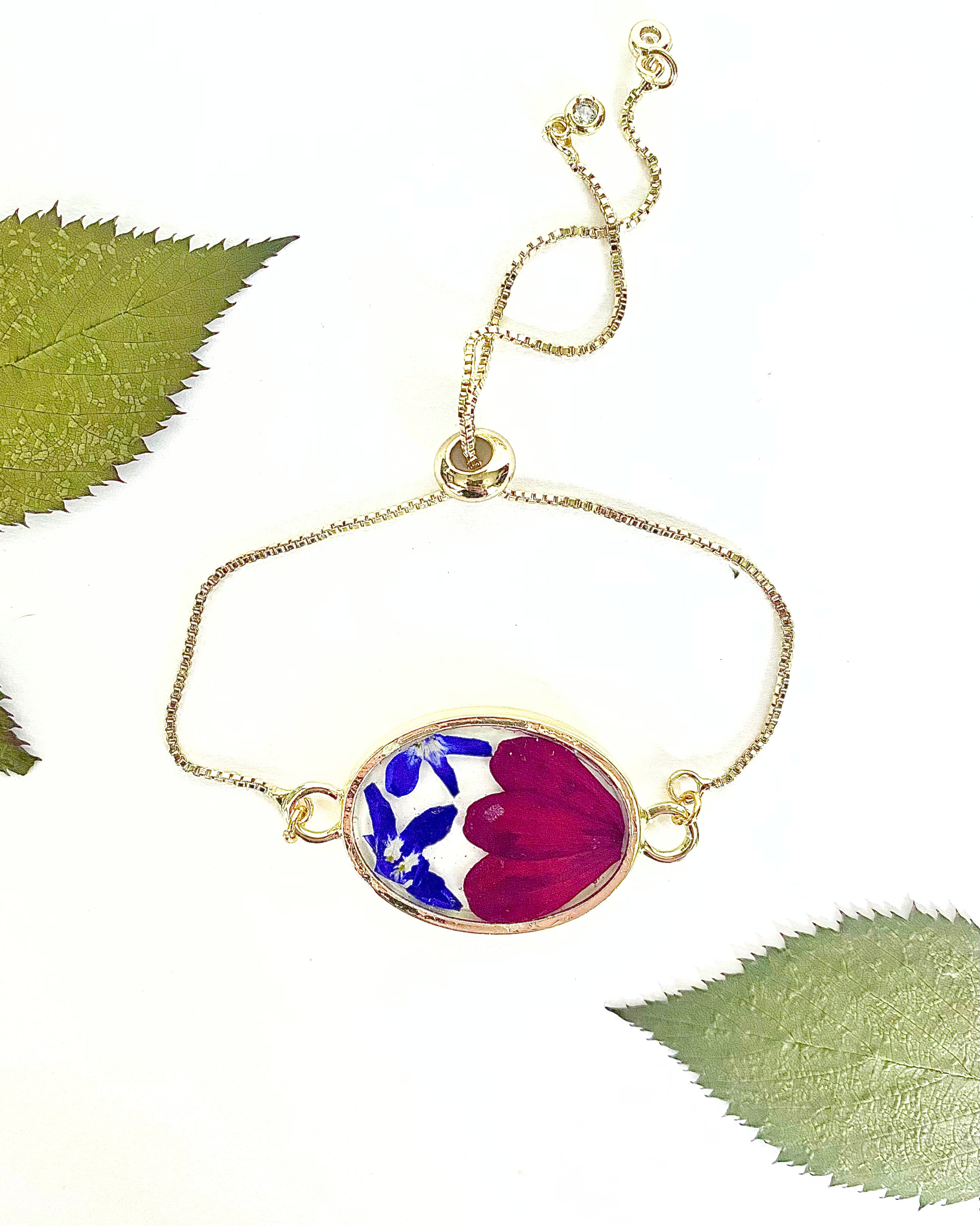 A delicate gold chain bracelet with an oval pendant containing pressed flower petals in blue, white, and red, displayed on a white background with decorative green leaves.