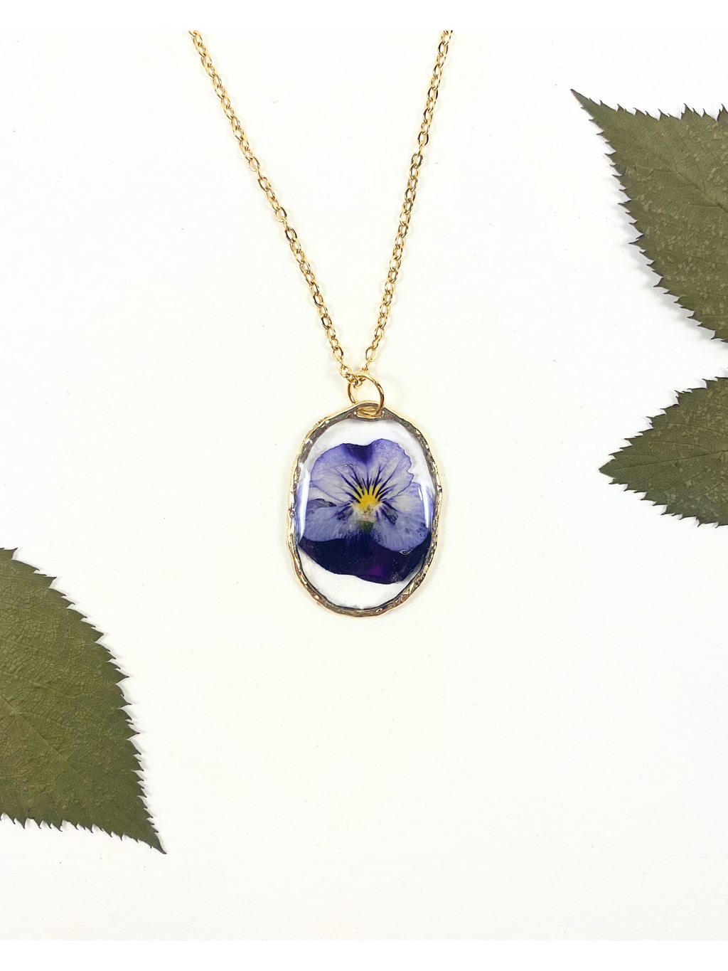 A gold chain necklace with an oval pendant featuring a preserved blue flower, styled on a white background with green leaves in the corners.