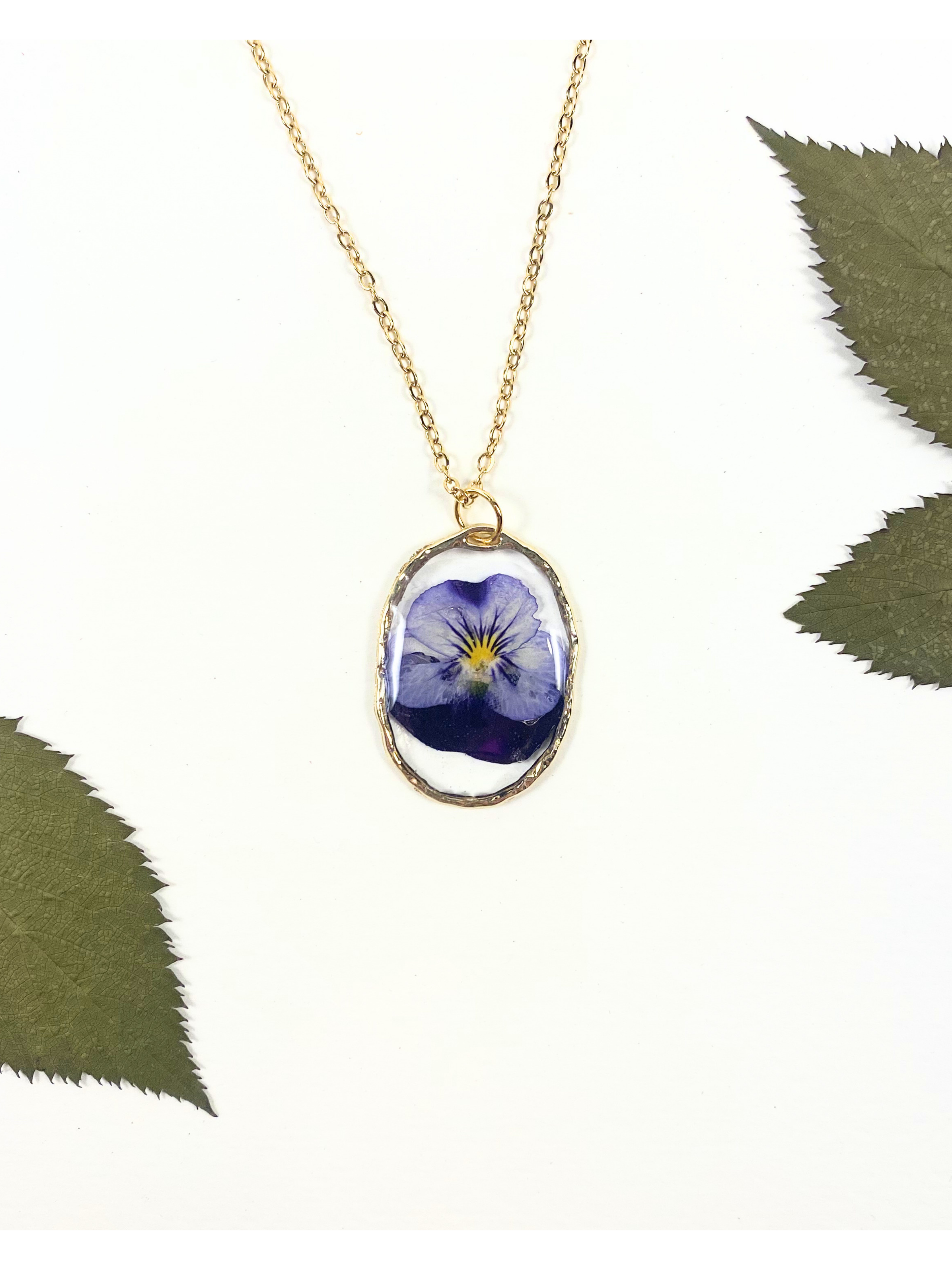 A gold chain necklace with an oval pendant featuring a preserved blue flower, styled on a white background with green leaves in the corners.
