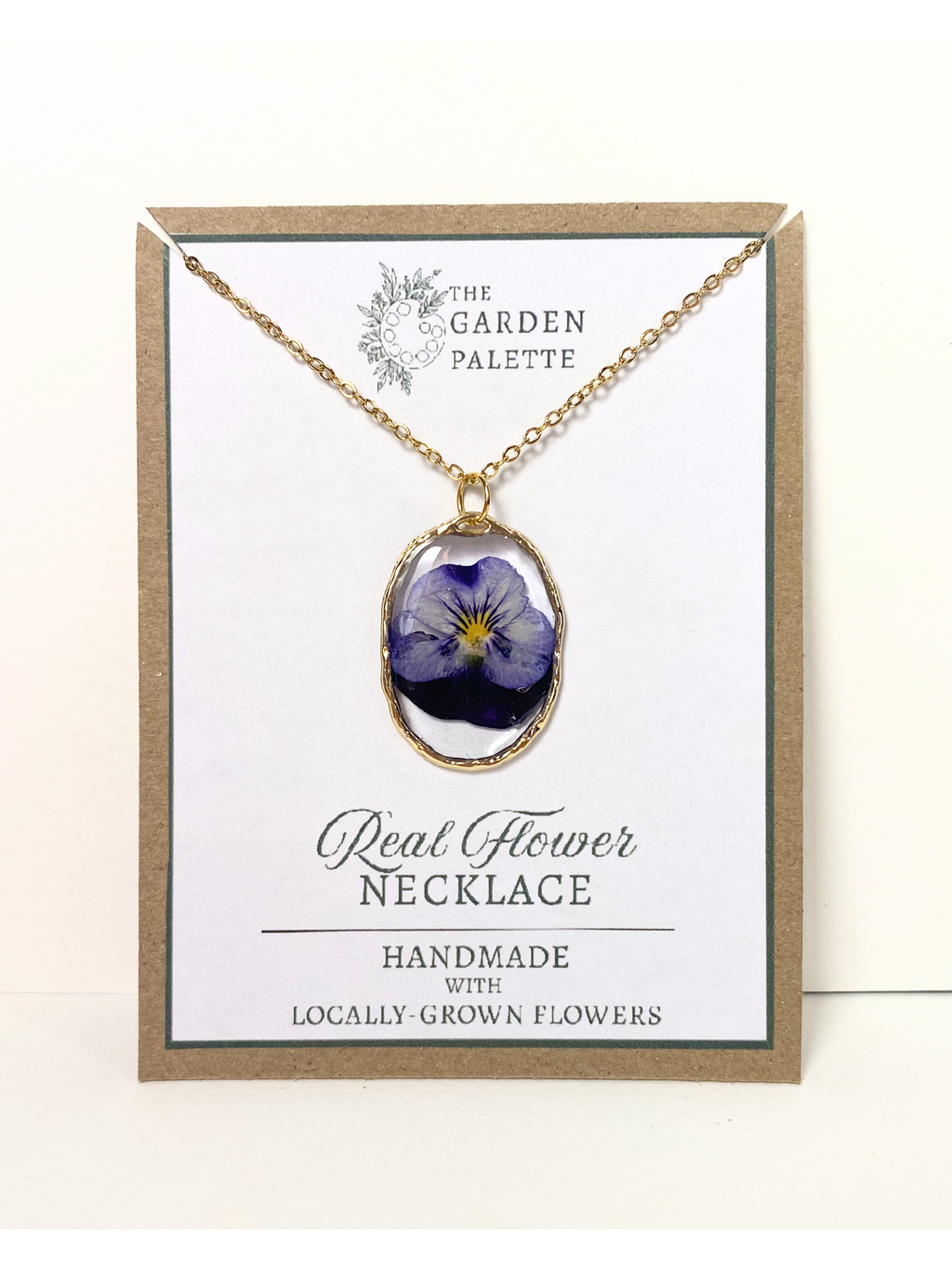 A gold-tone necklace with a real blue pansy flower pendant, displayed on a white card inside a kraft paper frame. The card reads 'The Garden Palette' and 'Real Flower Necklace'.