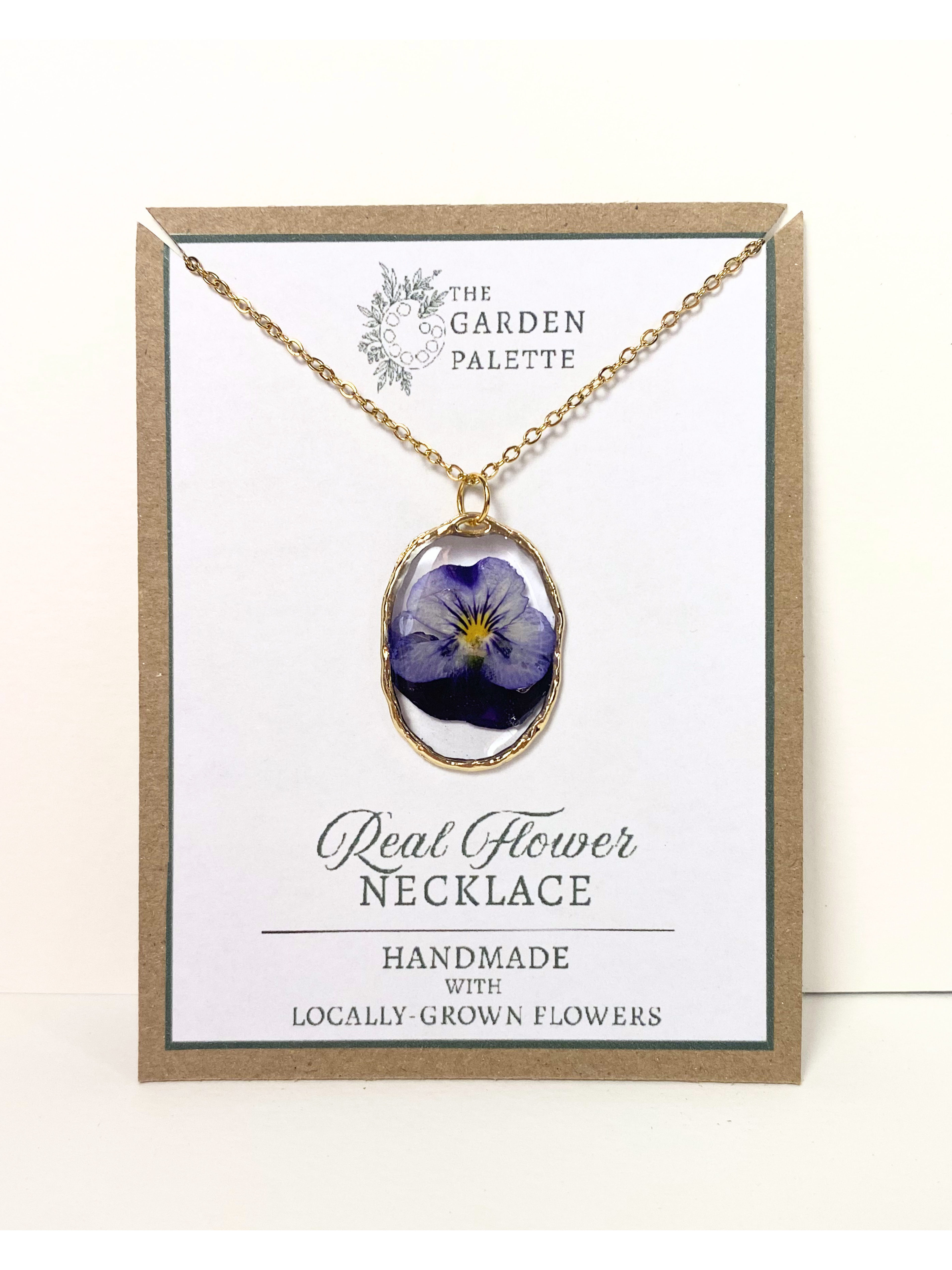 A gold-tone necklace with a real blue pansy flower pendant, displayed on a white card inside a kraft paper frame. The card reads 'The Garden Palette' and 'Real Flower Necklace'.
